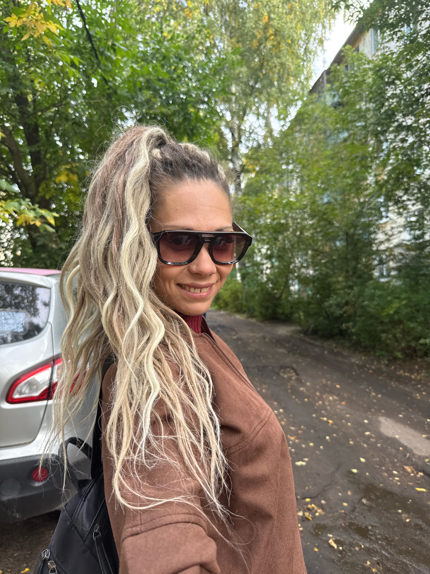 A blonde woman with long, wavy hair pulled into a high ponytail, wearing large dark sunglasses and a brown jacket over a red turtleneck, smiles at the camera. She is standing outdoors next to a silver car, possibly in a residential area. The background shows a dirt road with scattered autumn leaves and trees with green and yellow foliage, with a multi-story building visible in the distance. This setting is consistent with the location of Rusanovo, Russia, which is characterized by its natural surroundings and typical urban infrastructure. The lighting suggests it is daytime, and the overall atmosphere appears casual and relaxed.