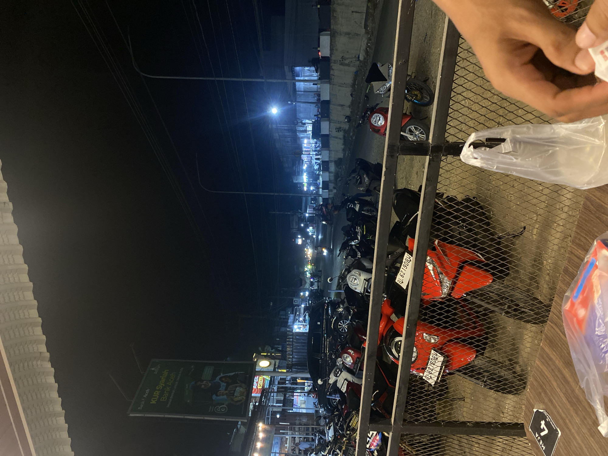 Here's a description of the image:
The photograph, taken at night in what appears to be Tanjongkapai, Indonesia, shows a street scene with parked motorcycles. The perspective is from an elevated position, possibly a balcony or upper floor of a building, looking out over the street. A metal fence or railing is in the foreground, partially obstructing the view.
Several motorcycles of varying models and colors are parked along the street. Some are oriented towards the viewer while others are positioned at an angle. License plates are visible on a few of the motorcycles, bearing the Indonesian license plate type. The vehicles seem to be closely packed together.
Across the street, there are buildings lit up with artificial lighting. A large billboard is visible on the left of the scene. It features advertising for "KUR Syariah Bank Aceh" with accompanying images and text, also showing "Apel Royal". Streetlights are present along the street, adding to the overall illumination.
In the upper right corner of the image, a person's hand is visible, holding a white plastic bag. The general lighting conditions are dim, but the light sources on the street create some contrast. The presence of overhead wires adds to the visual clutter of the scene.