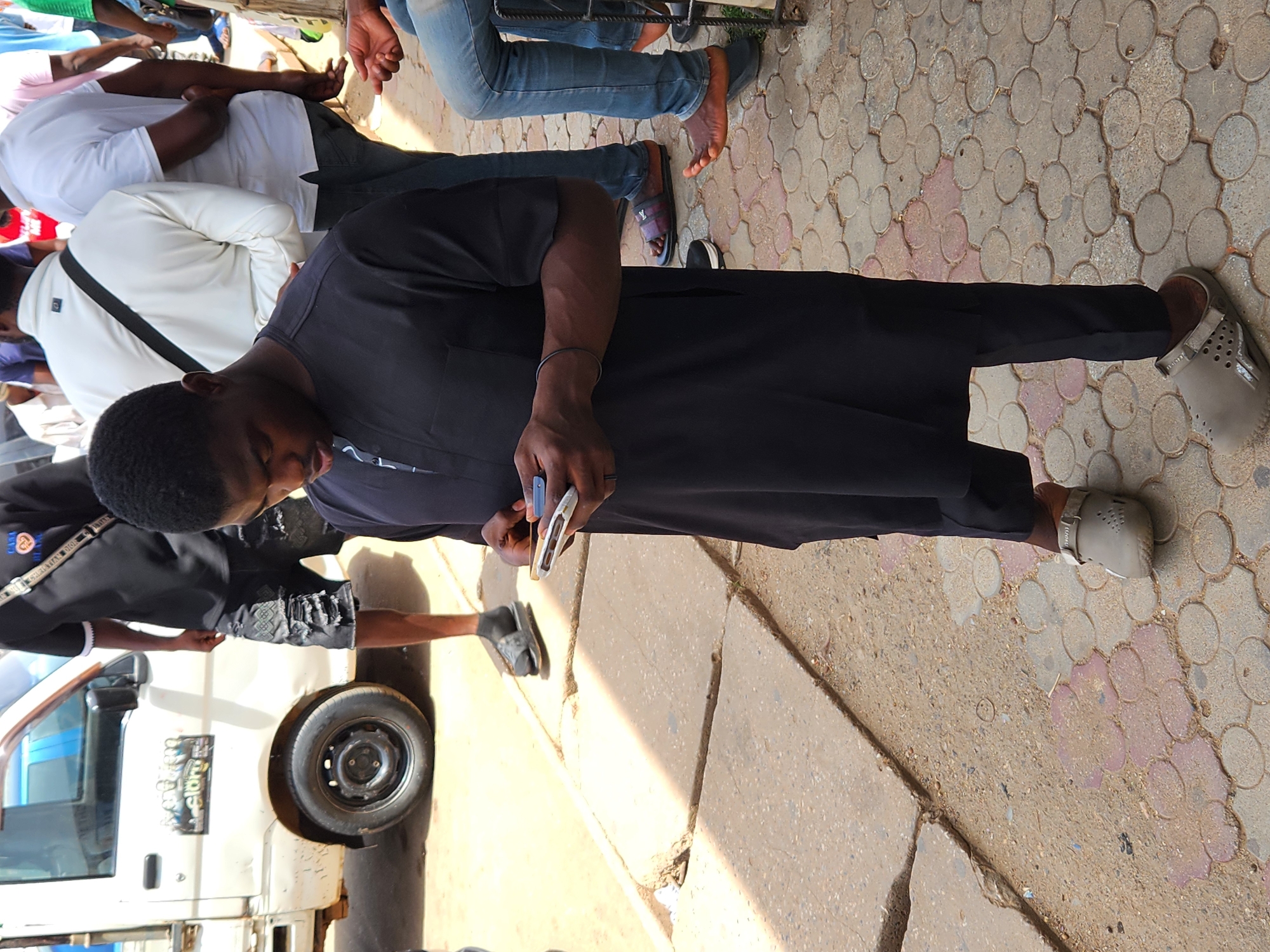The scene captures a street in Unknown, Unknown. A man wearing a black tunic and gray clogs is the primary focus, looking down at a blue object in his hands. He stands on a paved sidewalk with circular, interlocking stones, bordered by a concrete curb and an area of dirt or sand.
Several other people are partially visible. To the left of the man, a portion of a white van is present, along with a person in shorts and a black top standing near the driver's side door. Another person is seen wearing a white shirt. Some individuals wearing jeans and casual footwear are nearby as well. The overall setting appears to be an outdoor gathering.