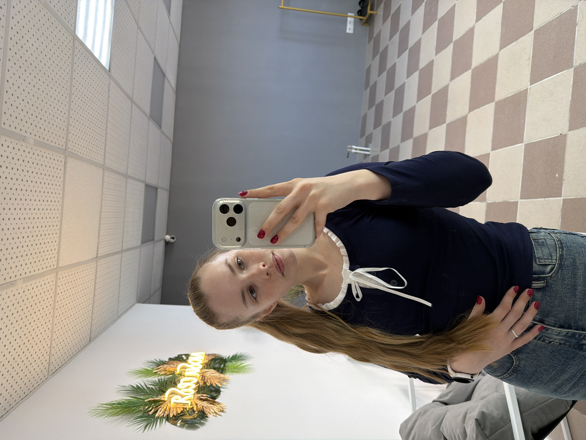 A fair-skinned woman is taking a selfie in an interior space with mirrored walls. She is wearing a navy blue top with a white ribbon accent and light blue jeans. Her long blonde hair is pulled to the side. She is holding a smartphone with a silver case, visible camera lenses, and red nail polish on her fingers. The background includes ceiling panels and a checkered floor. The mirror reflects a neon sign with the Russian name "Rostova". This scene is in Berezniki, Russia.