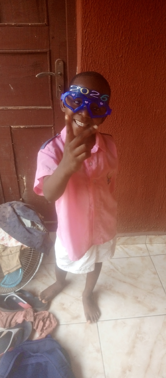 A child stands indoors, wearing heart-shaped glasses displaying "2026". The child is dressed in a pink short-sleeved shirt and white shorts, gesturing with a raised hand. The background includes a brown door on the left and a red wall on the right. Items like a fan, bag, and clothes are scattered on the tiled floor near the child's feet. The child is smiling.