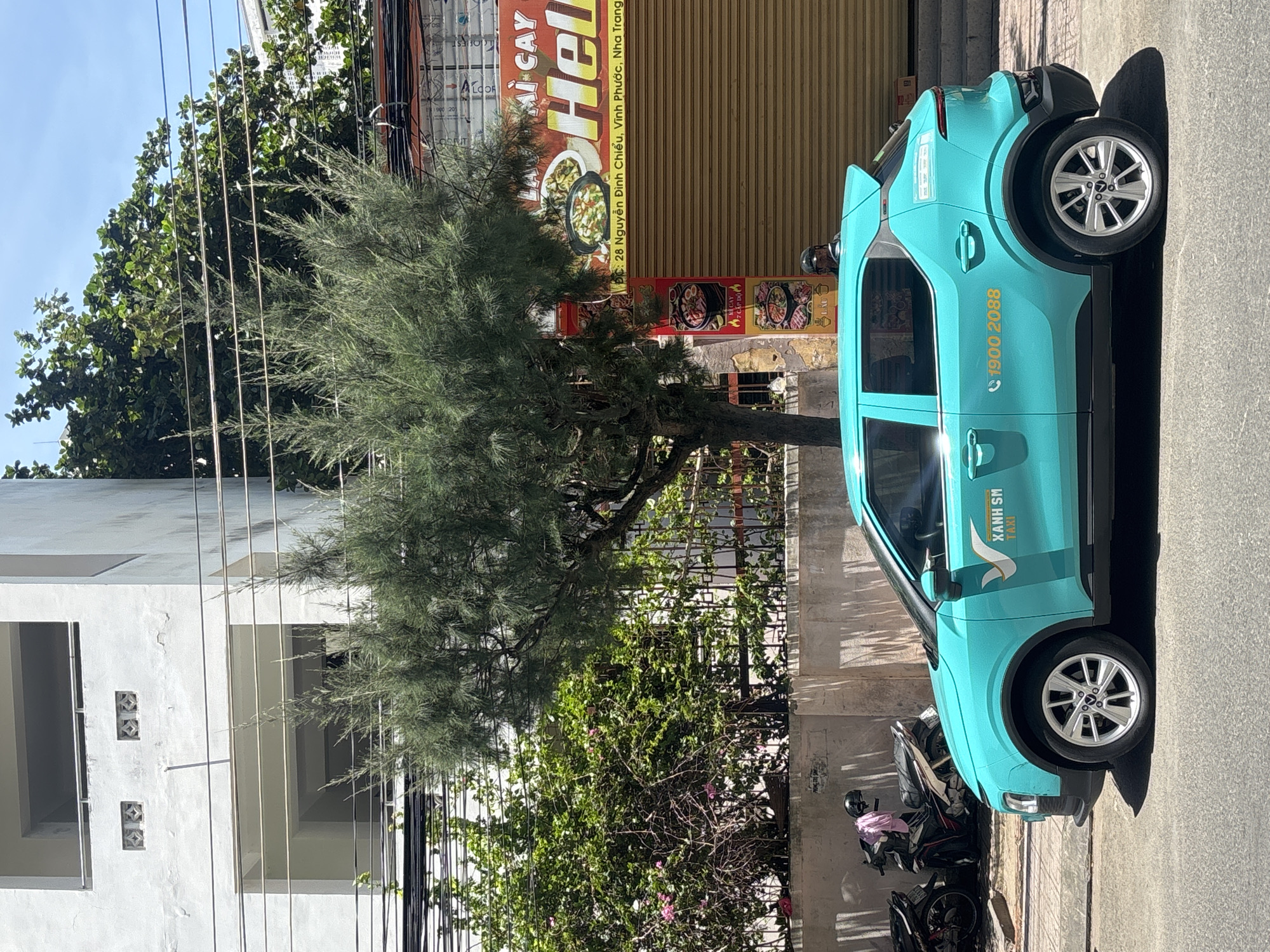 An eye-level medium shot captures a teal-colored taxi with "XANH SM TAXI" branding parked on a paved street. The taxi's side features a logo of a white bird and the phone number "1900 2088". Behind the taxi, a low, light-colored concrete wall runs horizontally, with lush green bushes and trees growing above it. To the right of the bushes, a shop with a large, rolled-down yellow shutter displays signage with Vietnamese text and images of food. Further behind, a multi-story white building with balconies and square window openings is visible, partially obscured by a tall, leafy green tree. In the background, a bright blue sky with thin white clouds stretches across the frame. To the left of the taxi, two motorcycles are parked against the wall.