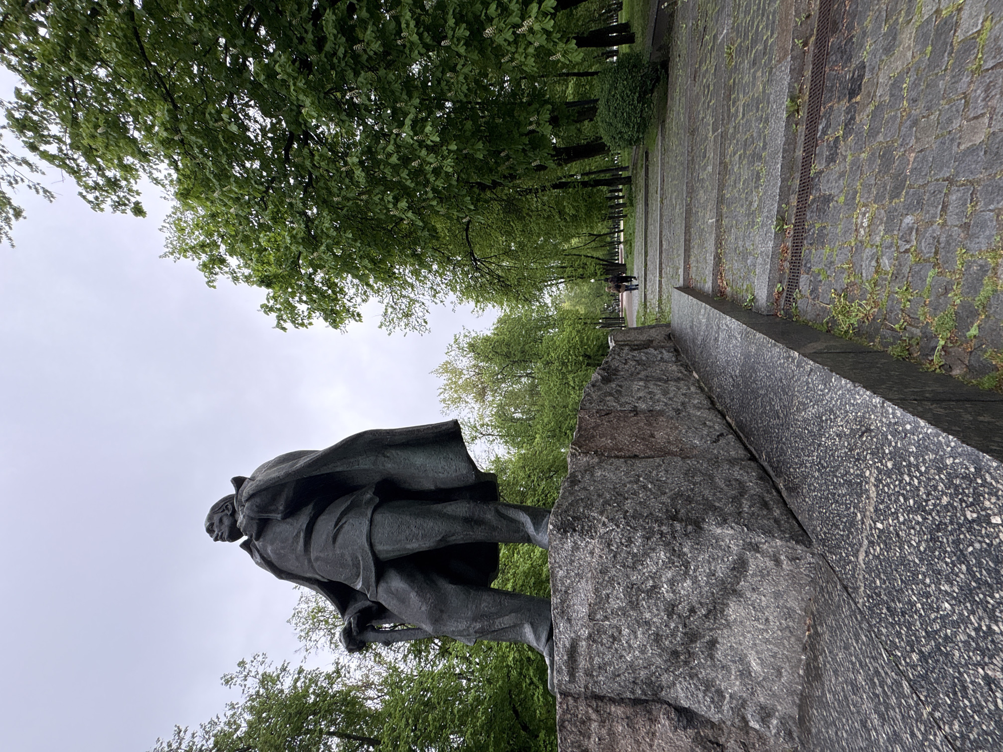 The image shows a statue of Maxim Gorky in Minsk, Belarus. The statue is a bronze figure standing on a stone pedestal. Gorky is depicted wearing a coat and hat. The background includes trees, paving stones, and a pathway with people walking in the distance. The sky is overcast.