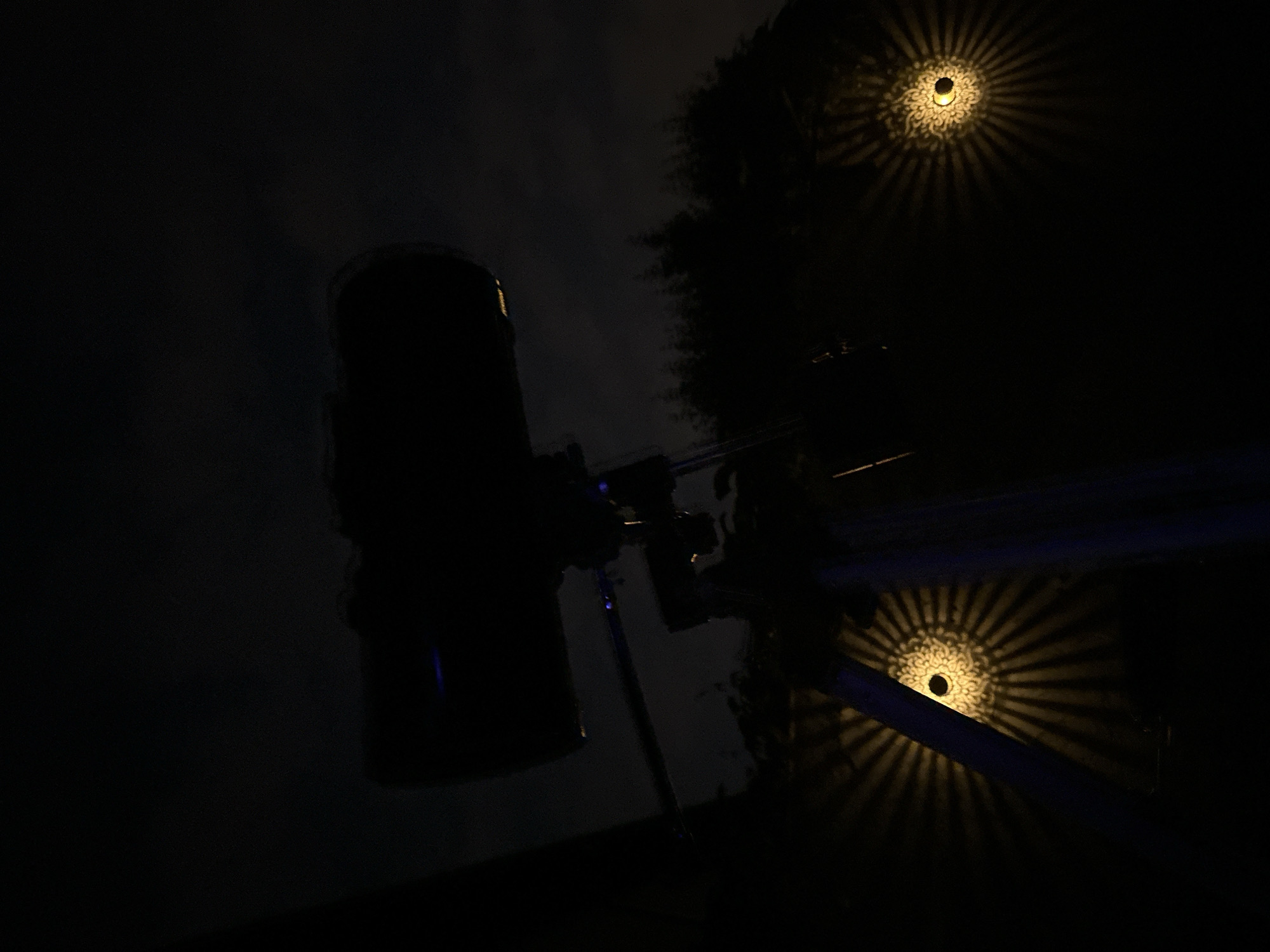 The image displays a nighttime outdoor scene in Villa Pasquali, Italy. A large astronomical telescope on a tripod is centrally positioned, silhouetted against a dark, partially clouded sky. Subtle blue light reflections are discernible on the telescope's barrel and mounting hardware. Two illuminated, wall-mounted light fixtures are present in the lower frame, one on the left and one on the right. Each fixture emits warm-toned light and projects a distinctive, intricate radial pattern onto an adjacent surface. The horizon in the mid-ground reveals a silhouette of coniferous trees. No individuals are visible.