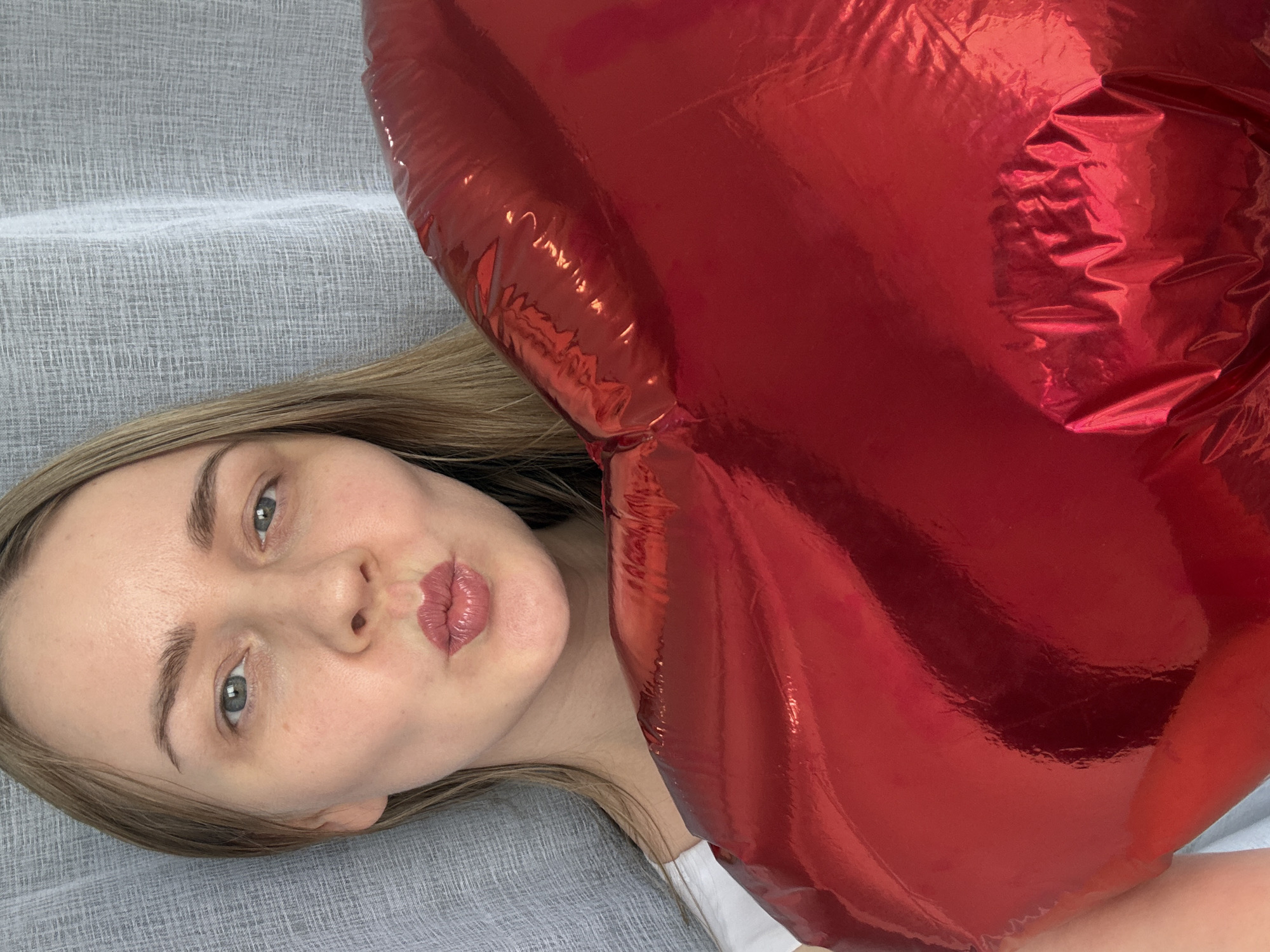 A woman lies on a gray textured surface, with a large, inflated red heart-shaped foil balloon obscuring part of her upper body. The woman has fair skin, light brown hair, and blue eyes. Her lips are pursed. She is wearing a white garment. The background appears to be a simple, indoor setting. The photograph was taken in Chaikovsky, Russia.