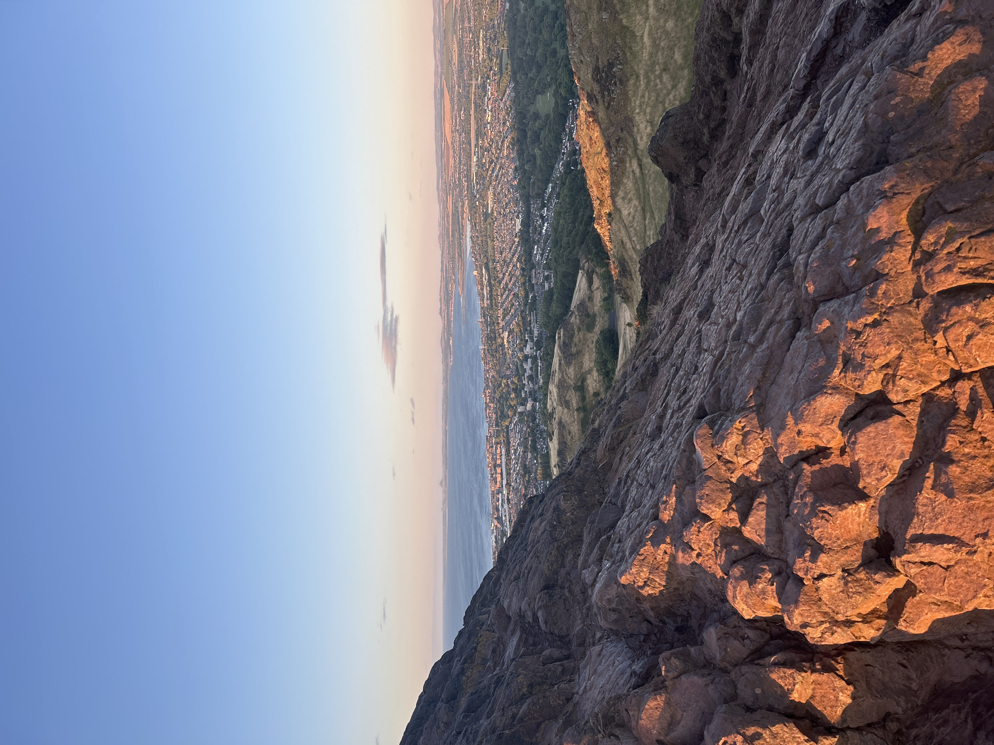 The image captures a panoramic view of the City of Edinburgh, Scotland, from an elevated vantage point, likely Arthur's Seat, at sunset. The foreground is dominated by rugged, reddish-brown volcanic rock formations, catching the warm, golden light of the setting sun, casting long shadows across the rough terrain.
Beyond the rocky foreground, the scene unfolds to reveal the sprawling urban landscape of Edinburgh. A vast expanse of the sea, the Firth of Forth, stretches across the mid-ground, its surface reflecting the soft hues of the twilight sky. Along the coastline, the city's buildings are densely packed, with a mix of architectural styles visible, suggesting a well-established urban center. Green spaces, including parks and possibly golf courses, are interspersed within the city, adding a touch of nature to the urban sprawl. Further in the distance, rolling hills and agricultural land frame the city, gradually fading into the horizon.
The sky above is a clear, serene gradient of pale blue, transitioning to softer, warmer tones near the horizon where the sun has set. A few wispy clouds are scattered across the sky, adding subtle texture to the vast expanse. There are no people or active elements depicted in the scene, giving it a tranquil and static quality. The overall impression is one of natural grandeur meeting urban development, bathed in the beautiful, soft light of a sunset.