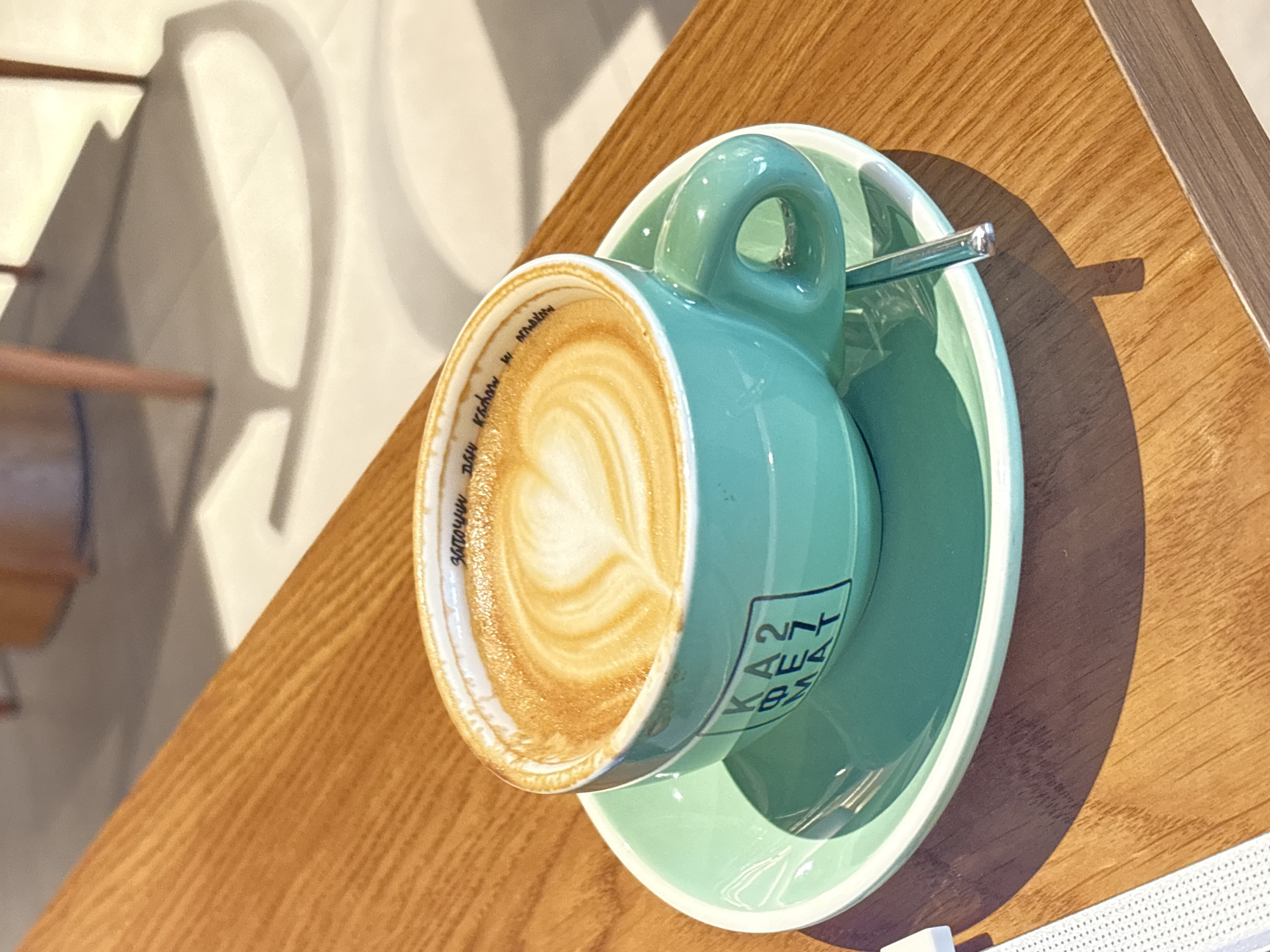 A turquoise coffee cup with latte art sits on a matching saucer on a wooden table, with a teaspoon partially visible. The cup's rim is labeled with text in Cyrillic script, including the phrase "заточим на ммакам". The cup also has printed text reading "KEAT 274 K85". The table appears to be indoors, with light suggesting a sunny day. The photograph was taken in Kragujevac, Serbia. A face mask is partially visible near the bottom right corner of the image.
