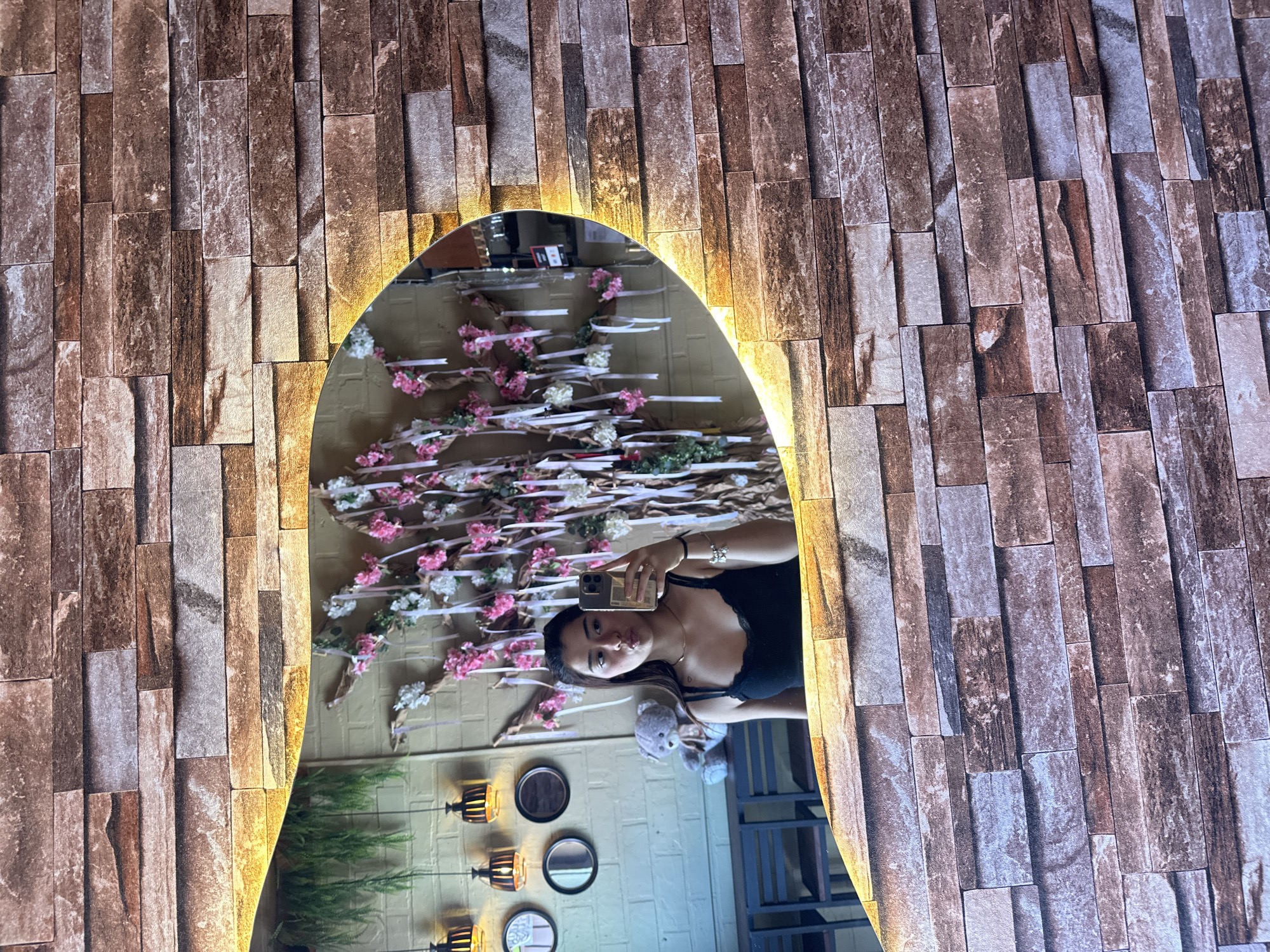 A woman is taking a selfie in a mirror with a decorative brick frame and internal lighting. The reflection shows a wall decorated with faux cherry blossom branches, framed by the mirror. Below, there are several small, round mirrors on the wall, as well as decorative lamps. A potted plant is also visible in the reflection. The woman is holding her phone to take the picture, and a teddy bear is visible in the reflection. The location is San Andrés Cholula, Mexico.
