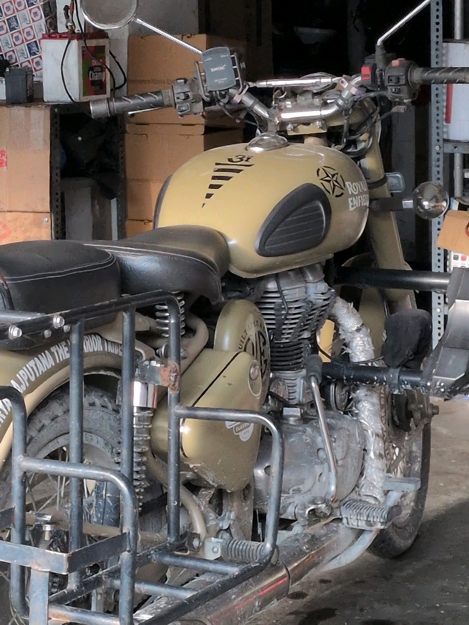 The image shows a motorcycle with a sidecar parked indoors, possibly in a garage or workshop. The motorcycle is a Royal Enfield, with "ROYAL ENFIELD" branding visible on the fuel tank. The fuel tank is a tan or light brown color, and the seat is black. The motorcycle has a sidecar attached, which appears to be made of metal tubing and also has a black seat. The motorcycle and sidecar are covered in a layer of dust and dirt, suggesting it may be old or has been used extensively. There are various boxes and shelves in the background, some containing what appear to be car batteries. The scene suggests a utilitarian setting, not a showroom. The location is not identifiable as Manali, India.