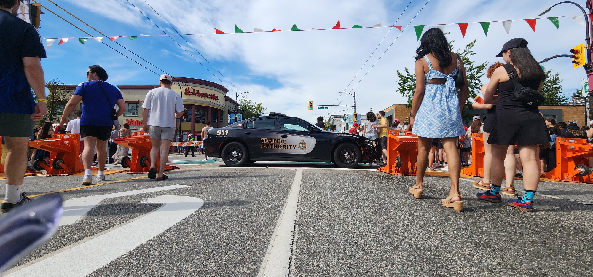 An eye-level, wide-angle shot depicts a street scene during what appears to be a daytime community event. In the foreground, the asphalt road displays white lane markings. In the midground, a black police car with "TRAFFIC AUTHORITY" and "911 EMERGENCY" text is positioned. Orange barricades are visible on either side of the road, some with wheels. Several people are present, including a man in a blue shirt and black shorts walking away from the camera, a man in a white t-shirt and grey shorts also walking away, and a woman in a blue patterned dress facing away from the camera. Other individuals are seen in smaller groups in the background, amidst buildings and trees. Overhead, a string of triangular flags in red, white, and green stretches across the sky, which is blue with scattered clouds. Traffic lights and street signs are also visible in the background.