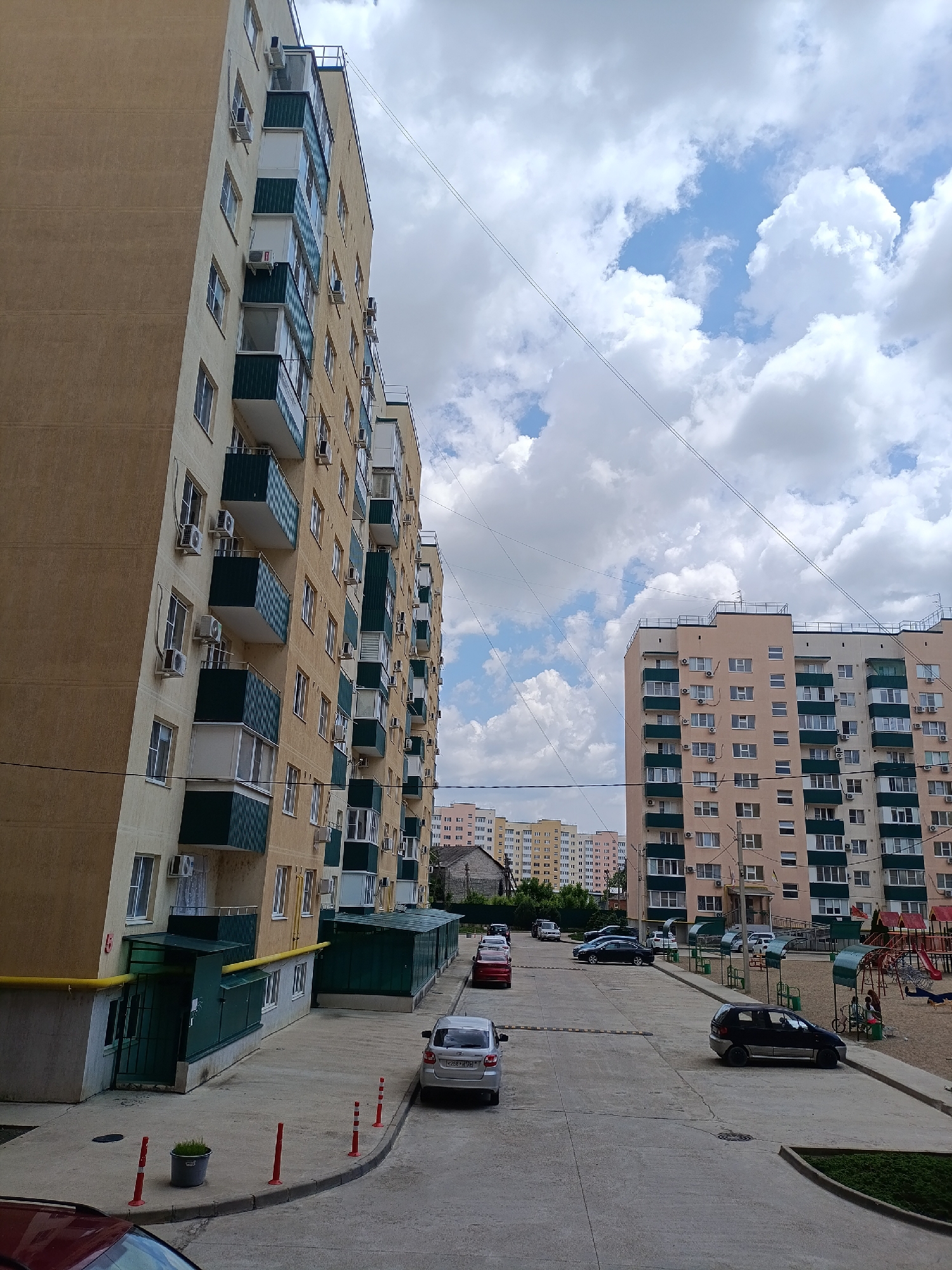 The image depicts a view within the city of Yablonovskiy, Russia, showcasing residential buildings under a sky with substantial cloud cover. The foreground features a paved area with parked cars, including a silver sedan and a dark minivan. Adjacent to the pavement is a curb lined with short red posts and a potted plant. To the left, a tall, multi-story residential building with a beige facade and balconies is visible. Across the street, a similar multi-story building with a peach-colored facade is present. Further in the background, additional apartment buildings can be seen. A children's playground with red play structures is visible on the right side of the image. Overhead, power lines stretch across the sky.