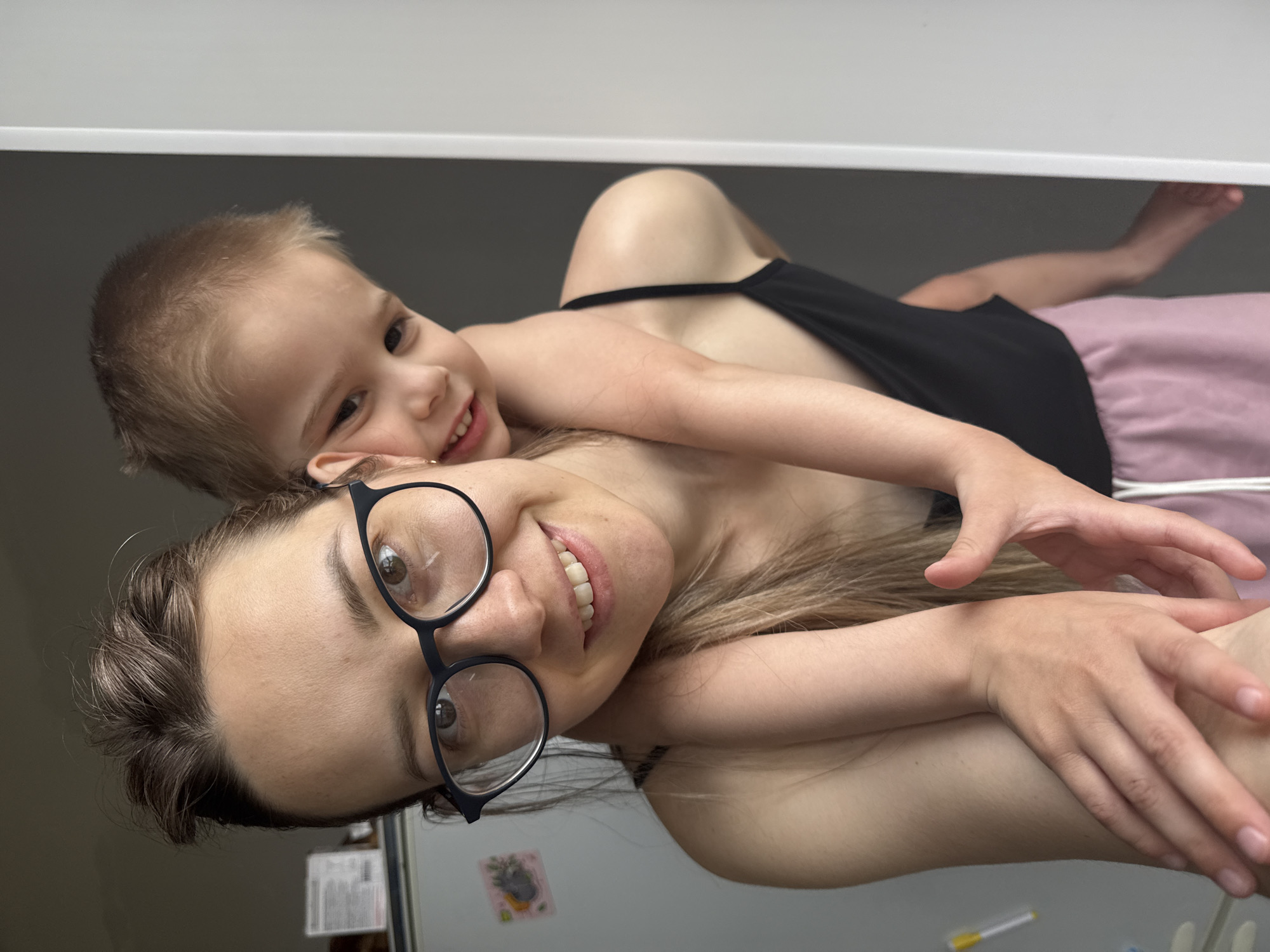A woman wearing glasses and a black tank top lies on her side, smiling at the camera. A young child with short blonde hair is lying on top of her, also smiling. The woman is wearing pink shorts. The background is a white wall and a glimpse of a refrigerator with a sticker. The location appears to be indoors, likely a domestic setting. It is impossible to determine whether this is in Chaikovsky, Russia, without additional information.