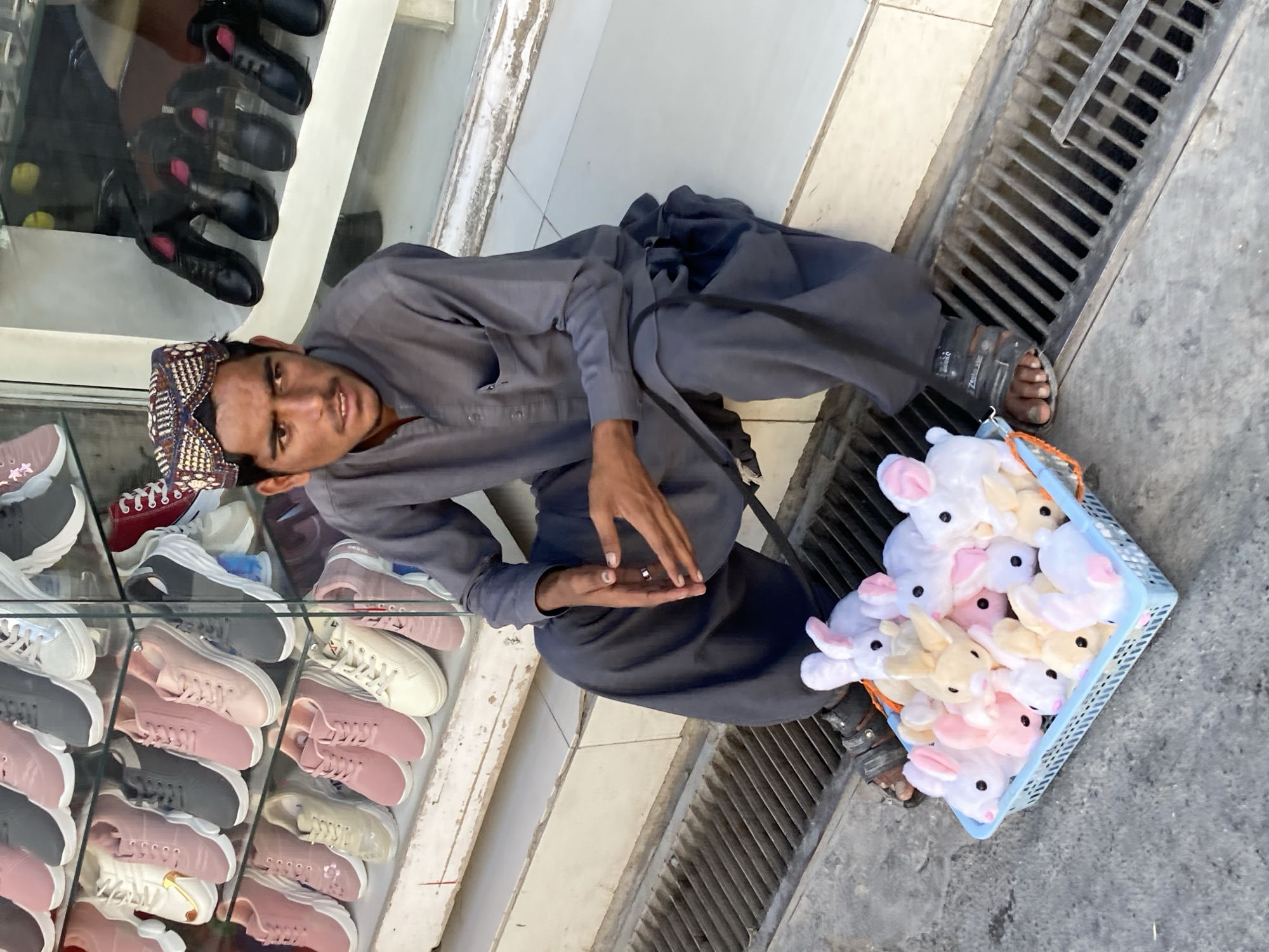 A man in a traditional Pakistani dress and cap kneels on the ground next to a drain. He is beside a shoe display case with mostly pink and grey sneakers. He holds up his hands, looking at the camera. A blue plastic basket filled with stuffed bunny toys sits beside him. The scene is set on a city street in Quetta City Tehsil, Pakistan, evident from the traditional clothing and urban setting.