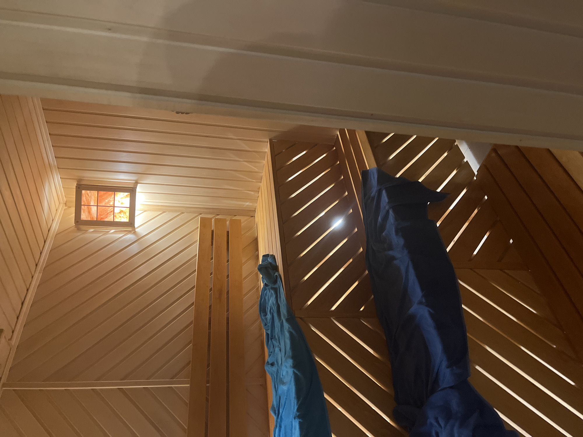 The image depicts an interior view with wooden planking on the ceiling and walls. A small, square window with a reddish glow is visible on the left wall. Two draped objects, one blue and one bluish-green, are suspended from the walls on the right side of the frame. The lighting appears to be a mix of natural light from the window and artificial light within the space. The diagonal placement of the planks on the right creates a pattern of light and shadow. The exact location in Новая Чернушка, Russia, is undeterminable from the visual information.