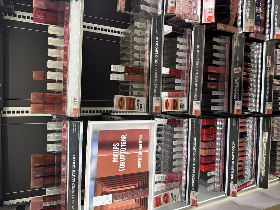 This image depicts a brightly lit retail cosmetics display, specifically featuring Maybelline lip products, within a store located in New York City, United States of America. The display consists of black shelving units with white vertical slotted tracks, supporting multiple clear plastic tiered trays. Various Maybelline lip products, including "24HR Blurred Matte Color," "Super Stay Matte Ink," "Longwear Matte Liquid Lipcolor," "Longwear Vinyl Liquid Lipcolor," "Longwear Matte Crayon," and "Creamy Lipstick," are arranged in organized rows. Product colors are predominantly shades of red, brown, and pink. A prominent sign on the upper left promotes "INK LIPS FOR UP TO 16HR. SUPER STAY MATTE INK." Smaller promotional cards feature images of lips and text such as "TRANSFER-PROOF." Price tags are affixed to shelves, indicating prices such as $14.49, $15.99, and $10.99, with some items displaying crossed-out original prices. Several product slots are empty across the display. No individuals are visible within the frame; the scene is static.