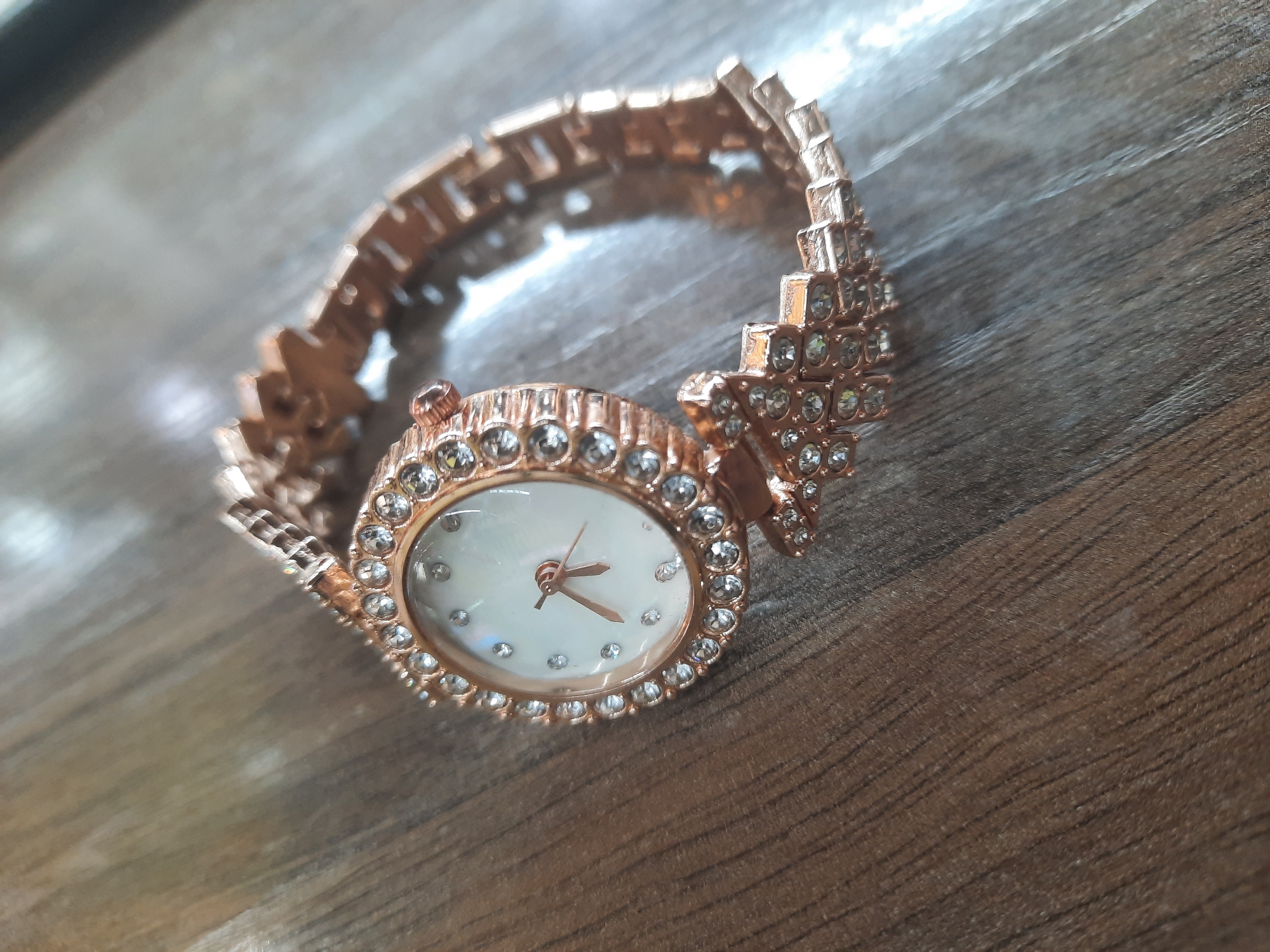 A close-up, elevated shot captures a decorative, rose gold-colored wristwatch resting on a textured wooden surface. The watch, likely designed for women, features an oval-shaped face with a pearlescent white dial, accentuated by delicate rose gold-toned hour and minute hands. Instead of numerals, small, sparkling clear gemstones mark the hours around the dial.
The bezel surrounding the watch face is encrusted with a continuous row of these clear, round gemstones, adding to its opulent appearance. A small, fluted crown is visible on the right side of the watch case. The watch strap, also in a rose gold tone, is intricately designed with multiple segments. The segments closest to the watch face are significantly wider and feature several rows of clear gemstones, forming a star-like or wing-like pattern that tapers into a more standard, though still ornate, link bracelet.
The wooden surface beneath the watch displays a prominent grain pattern with varying shades of brown, ranging from light beige to deeper, richer tones, suggesting an indoor setting. The lighting is soft and even, highlighting the metallic sheen of the watch and the sparkle of its gemstones without harsh shadows. No people are visible, and no activity is taking place; the watch is simply displayed. There is no visible text on the watch face or any other part of the item.