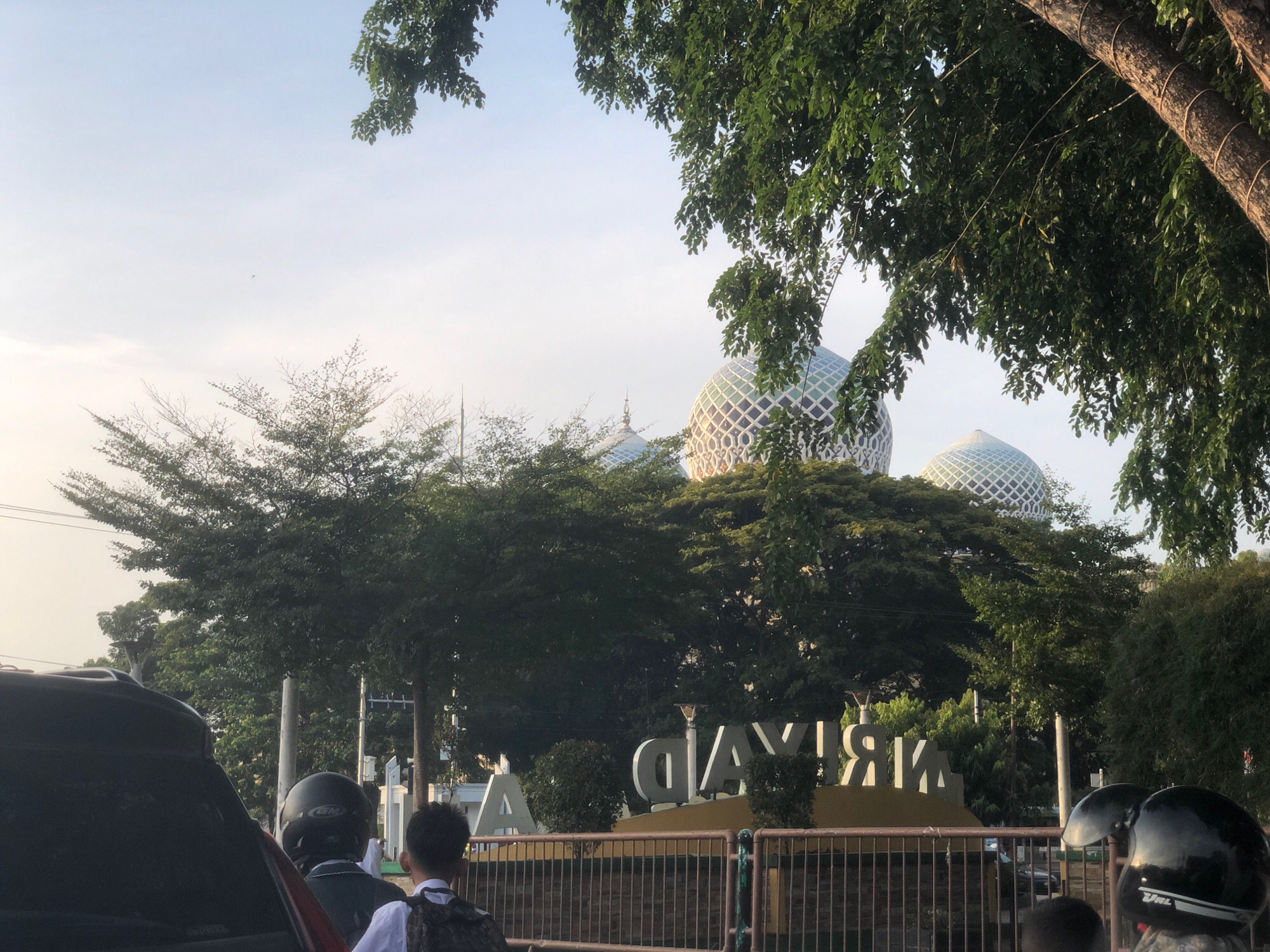 The image depicts a street scene in Lhokseumawe, Indonesia, featuring a prominent sign with the city's name, "LHOKSEUMAWE," rendered in large, white, three-dimensional letters against a yellow background. The sign is partially obscured by trees and fencing. Behind the sign, the distinctive domes of a mosque are visible, characterized by their intricate, patterned design, suggesting the Baiturrahman Grand Mosque or a similar significant religious structure in the city.
In the foreground, several people and vehicles are present, indicating a busy urban environment. A dark-colored car is partially visible on the left, with a person wearing a black helmet seated on a motorcycle behind it. Further to the right, another individual, appearing to be a student, is visible from behind, wearing a backpack. A metal fence runs across the lower portion of the image, separating the street from the area with the sign and mosque. Lush green trees are abundant throughout the scene, framing the view of the mosque and the city's signage. The sky is light and appears to be either early morning or late afternoon, given the soft illumination.