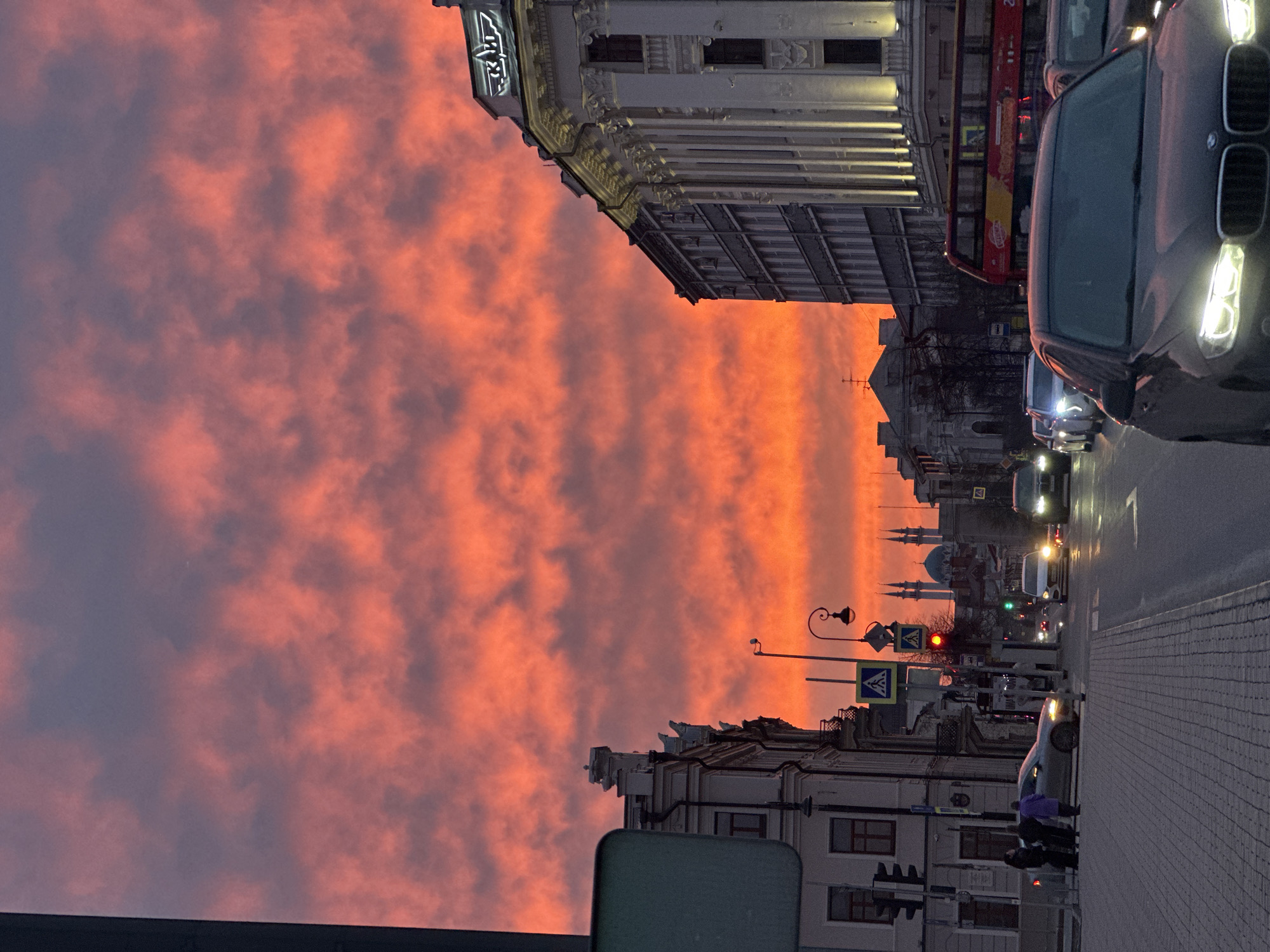 The image depicts a street scene in Kazan, Russia at dusk. The sky is ablaze with orange and pink clouds. A line of sight reveals the silhouette of the Qolşärif Mosque with its blue domes and minarets in the distance. The street is lined with buildings, including a tall, ornate structure on the upper left. A red double-decker bus can be seen behind a dark-colored SUV with illuminated headlights. Several other cars are present. Pedestrians are visible on the sidewalk near a traffic light and a crosswalk sign. The street is paved with gray brick or stone.