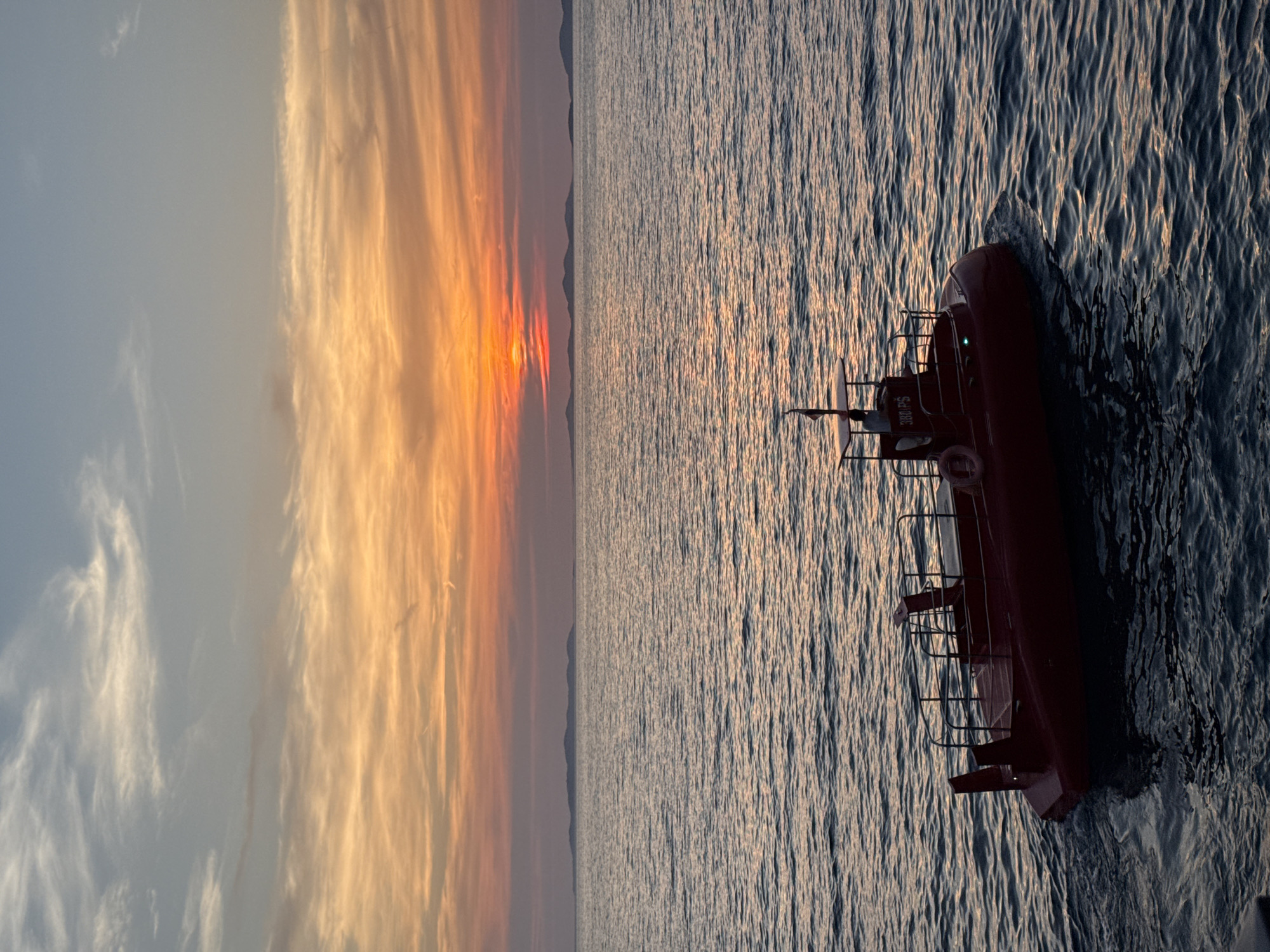 A red submarine is on the water with the sunset in the background. The sky is painted with shades of orange and pink from the setting sun. The water is dark blue with small waves. There are islands in the distance on the horizon. The submarine has a small cabin and railings around the top. There appears to be a person inside the cabin.