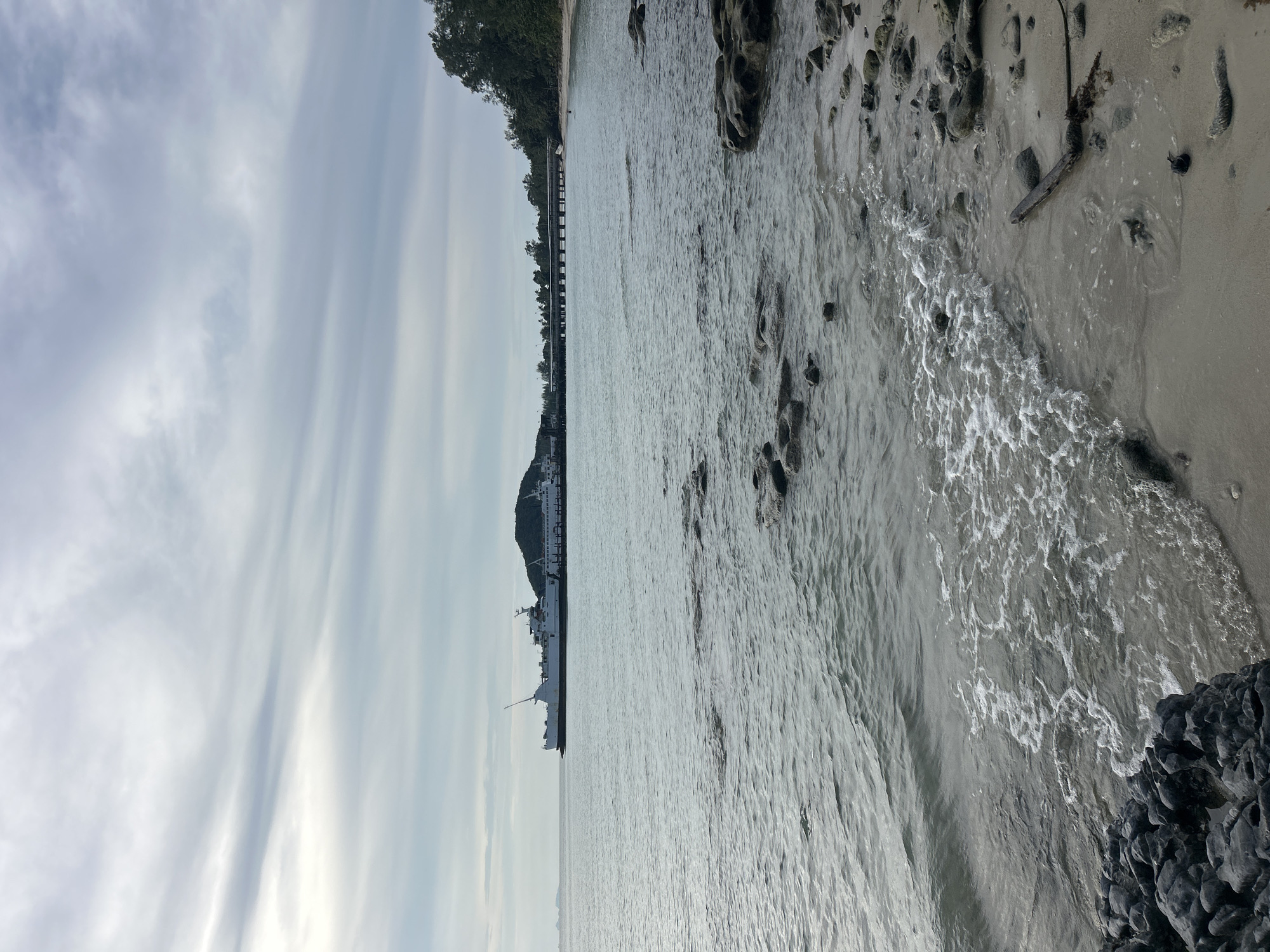 An overcast day at a ferry terminal in the Ko Samui District of Thailand. A large white ferry is docked at a pier, with a smaller ferry visible behind it. The pier extends into the calm, grey water. On the right side of the image, a line of trees marks the edge of a sandy beach. The foreground shows a rocky shore with waves gently lapping at the sand. The sky is filled with soft, grey clouds. There are no visible people in the image.
