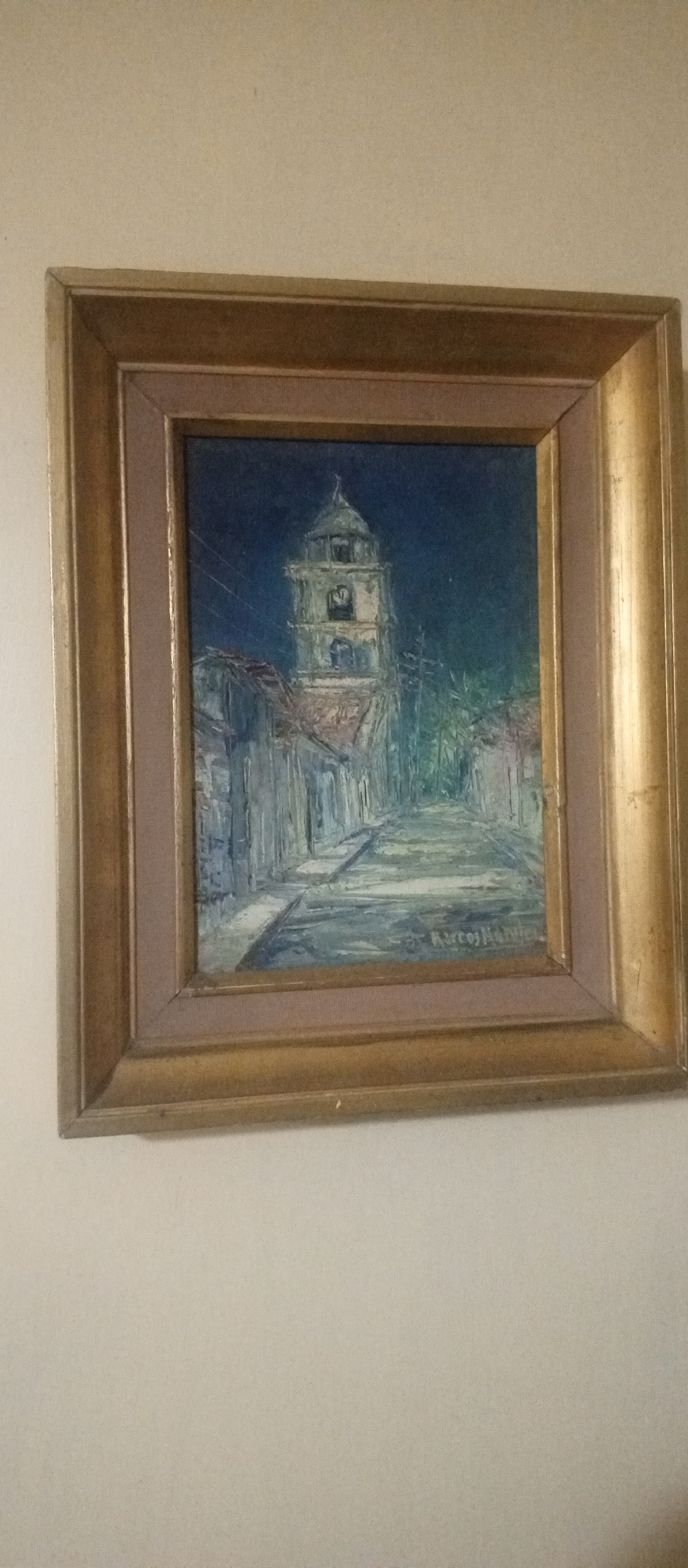 A framed oil painting depicting a street scene in Tinaquillo, Venezuela, is mounted on a wall. The painting's focal point is a church bell tower rising above a row of buildings lining the street. The buildings are painted in muted tones, while the bell tower is rendered in lighter colors. The street leads toward the tower, giving the viewer a sense of perspective. Overhead electric or telephone lines are also visible. The painting is signed "Marcos Mariel" in the lower right corner. The frame is ornate, featuring gold trim and a light brown inner border. The wall is plain, and light in color.