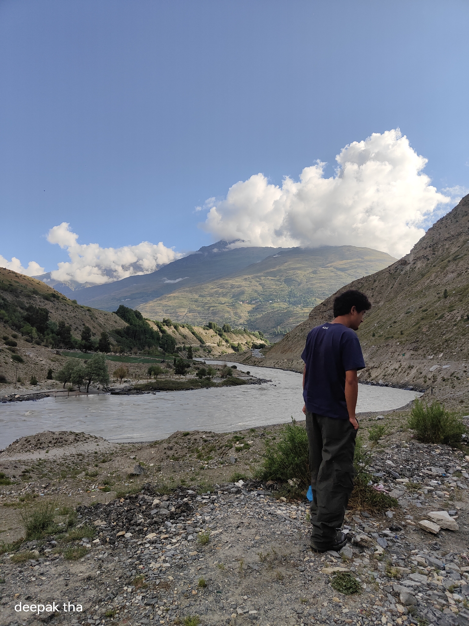 A person stands with their back to the camera, looking out at a wide, calm river flowing through a valley. The person is wearing a dark blue t-shirt and dark green pants. The landscape is mountainous, with dry, rocky slopes on the right and gentler, vegetated slopes on the left. Trees are visible on the left bank of the river, and there are fields or terraced areas further up the mountain. The sky is blue with scattered white clouds, and the sun is casting shadows, suggesting it is daytime. The foreground is a rocky, gravelly path. The text "deepak tha" is visible in the bottom left corner of the image.