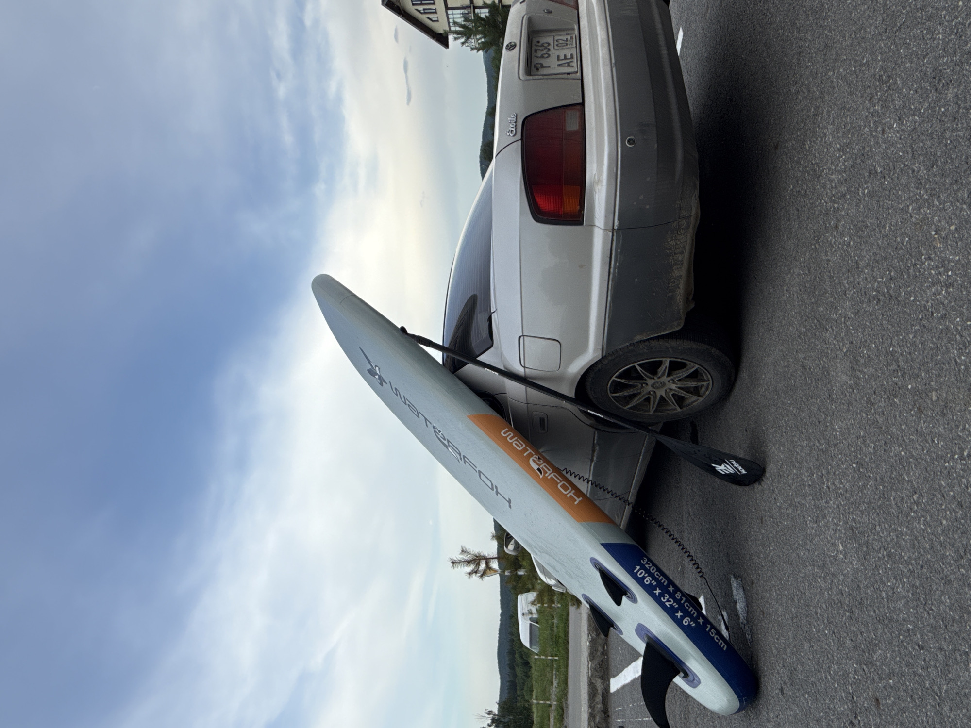 An eye-level, outdoor, slightly high-angle shot captures a silver sedan parked on an asphalt surface. A light blue and orange inflatable stand-up paddleboard is propped against the trunk of the car, resting diagonally on the rear quarter panel and the rear bumper. A black paddle with "Kaitsu" printed on it lies on the asphalt next to the paddleboard. The car's rear license plate displays "P 636 AE 02". In the background, a white van is parked on a grassy verge, and a building with multiple windows and a dark roof is visible on the right. Trees and hills form the distant landscape under a partly cloudy sky. The scene appears to be located in Syrostan, Russia, given the license plate format and surrounding environment. There are no people visible in the image.