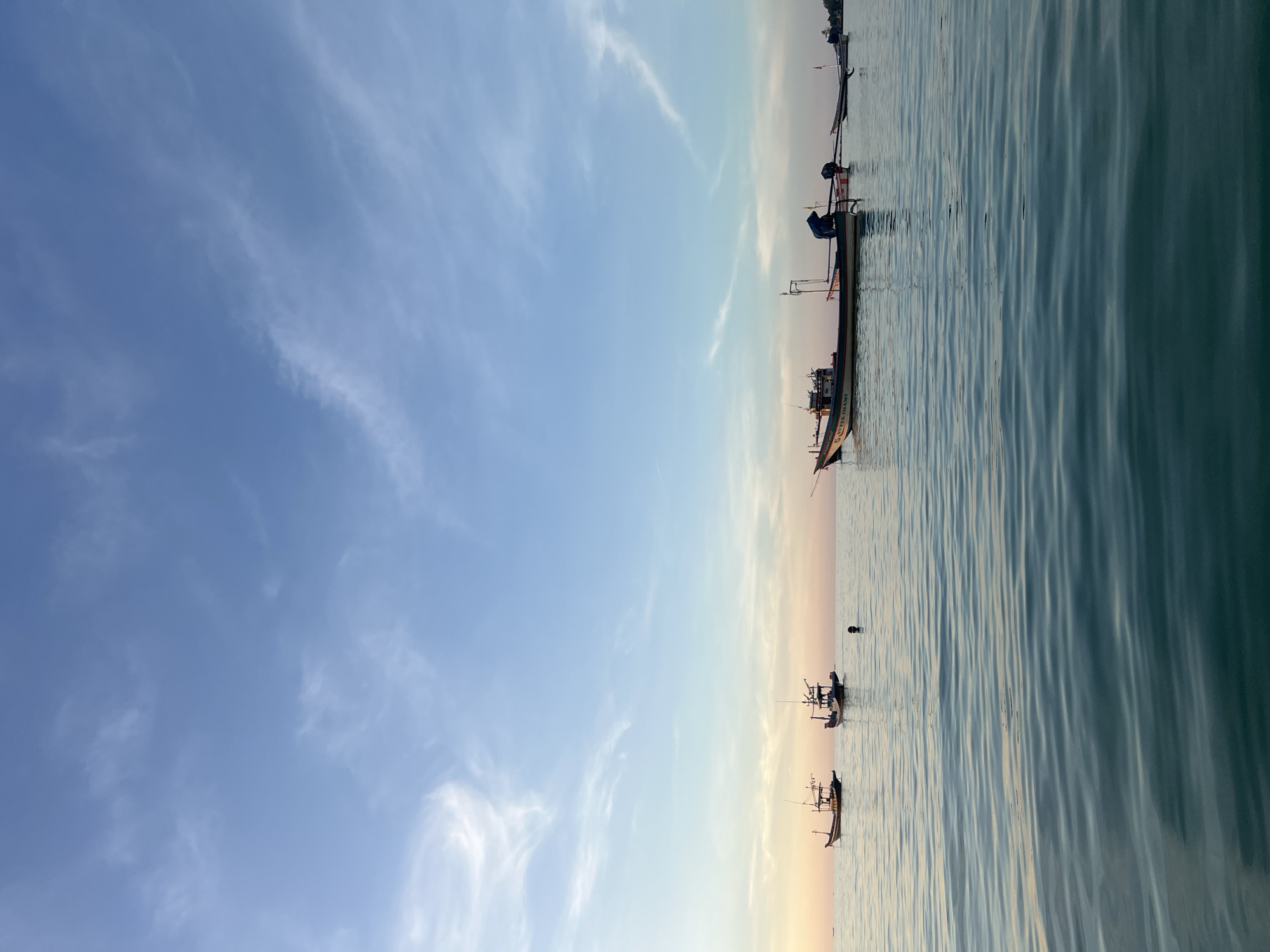 The image depicts a tranquil seascape with several fishing boats anchored on calm, rippling water under a pale blue sky with wispy white clouds. The sky transitions to a soft orange hue near the horizon, suggesting either sunrise or sunset. The scene appears to be set in the waters off the coast of ตำบลเกาะพะงัน, Thailand, given the characteristic style of the fishing vessels and the tropical sky.
In the foreground, the water's surface is a deep teal, reflecting the sky and creating a serene, undulating pattern. The middle ground features a cluster of fishing boats of varying sizes and designs. On the left, two smaller, darker boats are anchored relatively close together. Further to the right, a larger boat, predominantly white with colored stripes, is prominently positioned. It has a cabin structure and appears to be equipped for fishing. Beside this larger boat, a smaller, narrower boat is visible, with a person wearing a dark garment visible inside. To the far right, another slender boat is anchored.
In the distance, along the horizon, a faint landmass can be discerned, possibly the coastline or other islands. The overall atmosphere is peaceful and quiet, evoking a sense of calm before or after a day's fishing. The lighting is soft, indicative of the early morning or late evening hours.