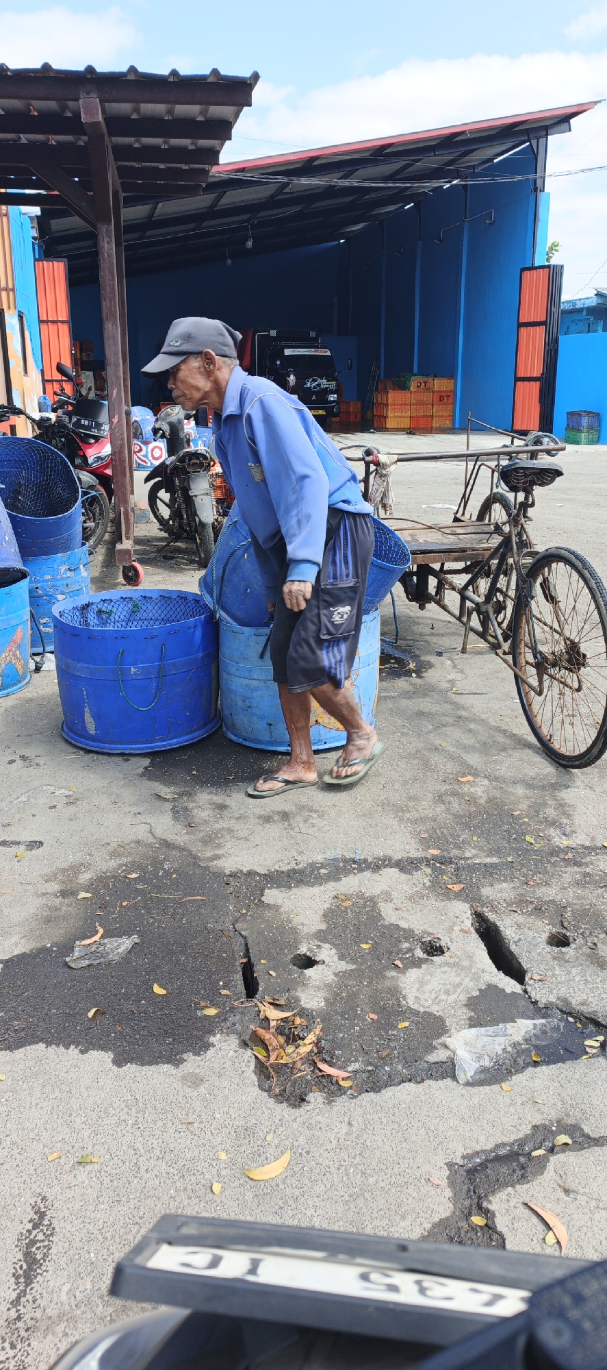 The image depicts an outdoor scene at what appears to be a fish market or a similar wholesale distribution area in Puger, Indonesia. A man, wearing a blue long-sleeved shirt, black shorts with blue stripes, a grey baseball cap, and flip-flops, is actively engaged in handling large blue plastic barrels. He is bent over, with his right hand near the rim of one of the barrels. These barrels, some with netting inside, are arranged on a cracked asphalt surface.

Adjacent to the barrels, a rusted, older model bicycle with a cargo carrier is parked. In the background, there is a covered structure with a corrugated roof supported by metal poles. Behind this structure, a black truck is visible, along with stacks of orange crates and blue and green plastic containers. Two motorcycles are also present, parked to the left of the man. The ground is wet in places and littered with fallen leaves. The sky above is partly cloudy. The overall atmosphere suggests a working environment, likely related to the seafood industry, common in coastal towns like Puger.