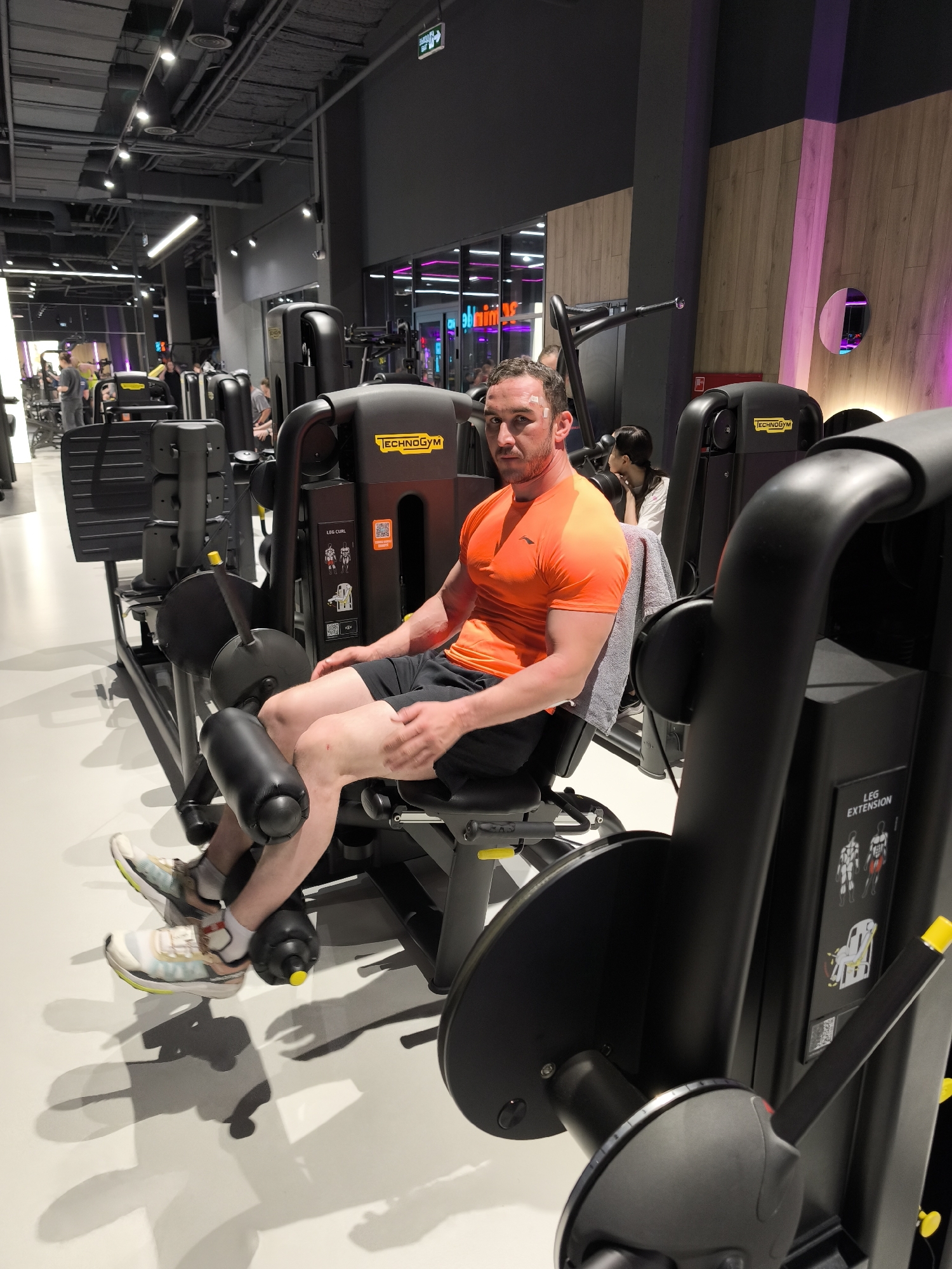 The image shows a man in an orange t-shirt sitting on a leg extension machine in a gym. He has a small bandage on his forehead and is wearing black shorts and athletic shoes. The leg extension machine is black with the brand name "Technogym" visible. Other gym equipment is visible in the background, along with other individuals working out. The setting appears to be a modern, well-lit gym facility. According to the image source, this photo was taken in Moscow, Russia.