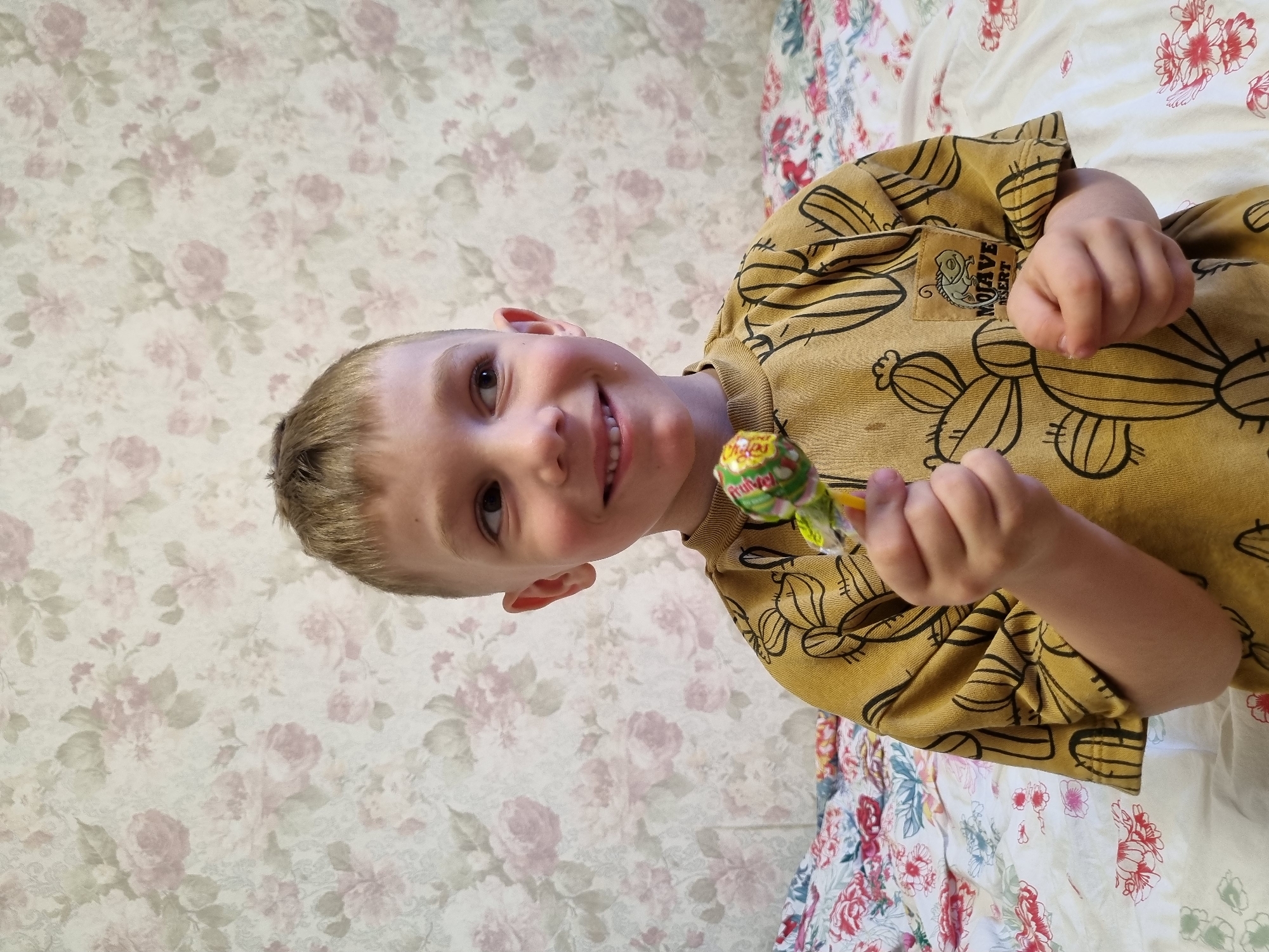 A young boy is holding a lollipop while smiling. He is wearing a gold shirt with a cactus print and the words "Mojave Desert" on the tag. The background is a floral wallpaper. The picture was taken indoors, most likely in a home in the city of Chaikovsky, Russia.