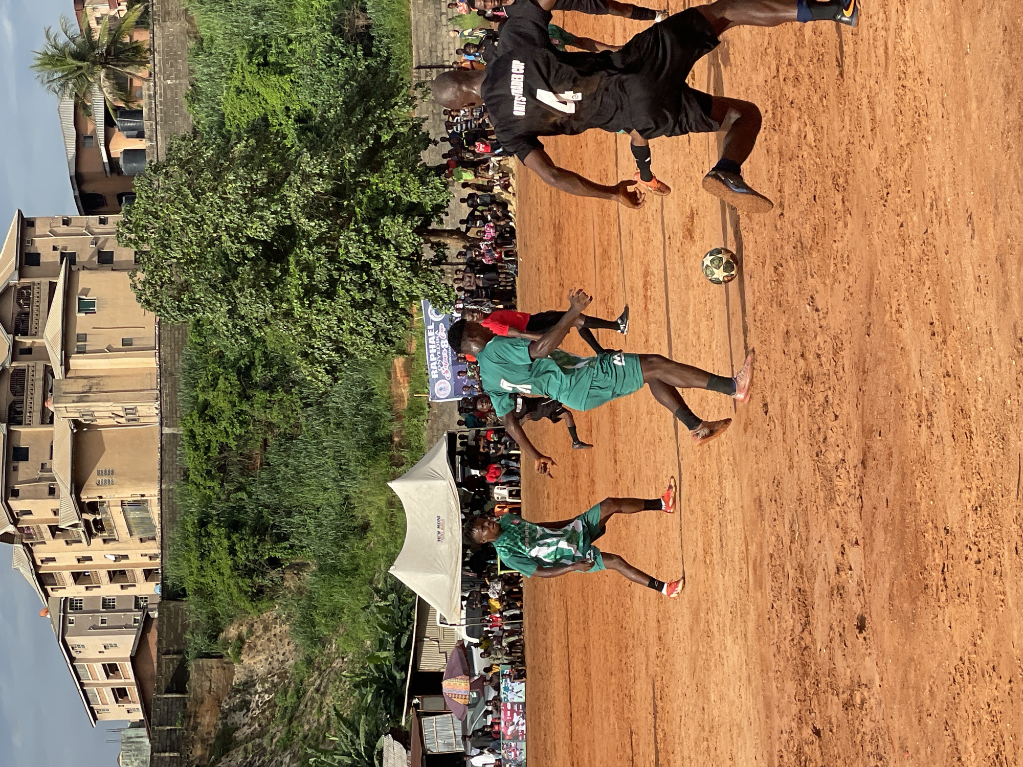 An outdoor soccer match is underway on a dirt field under a cloudy sky. Several players in green and black uniforms are engaged in the game. One player in a green and white striped jersey and shorts with orange cleats is in the foreground, dribbling a soccer ball. To his right, a player in a black jersey with the number 4 on the back and black shorts is running, with one leg extended towards the ball. Another player in a green jersey and shorts is further back on the left, also in motion. Spectators are gathered behind a makeshift fence to the left of the field, some seated under a white tent. In the background, multi-story buildings with beige and brown facades are visible on a hillside, surrounded by lush green vegetation. A tall tree stands to the right of the tent.