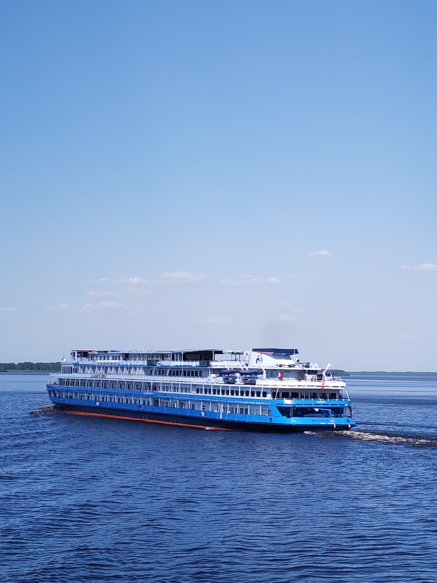 A medium shot captures a bright blue and white cruise ship sailing on a dark blue body of water, presumably in Selishchi, Russia. The ship has multiple decks and numerous windows. It leaves a white wake in its path. The sky above is mostly clear blue with a few scattered clouds. A distant shoreline with trees is visible in the background.