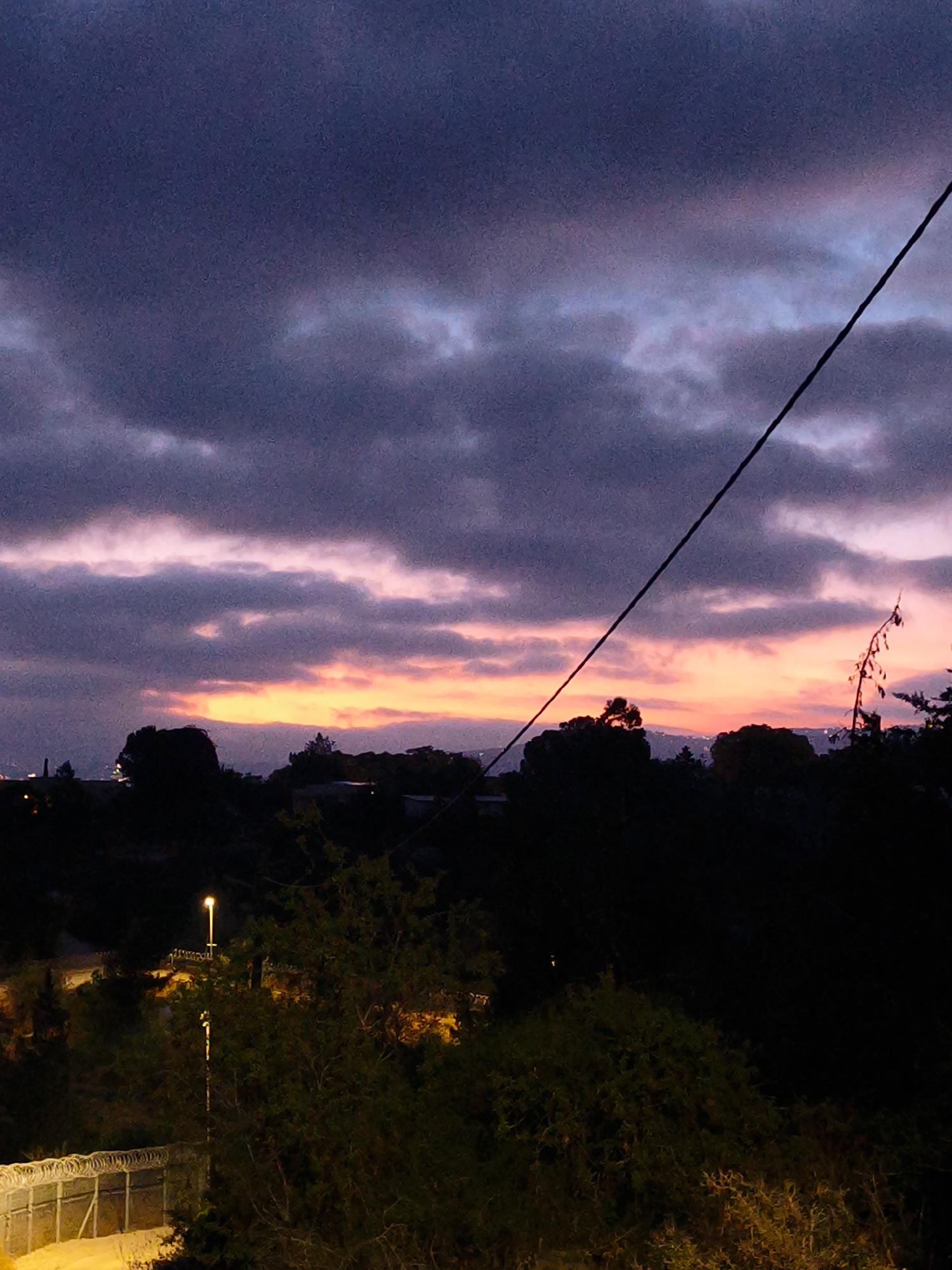 This image captures a twilight scene in the Lachish Regional Council, Israel. The sky is a dramatic display of dark purple clouds tinged with streaks of pink and orange from the setting sun. A solitary black wire bisects the sky diagonally. In the foreground, silhouetted trees and foliage create a dark, dense border. Further back, the faint glow of distant city lights is visible against the horizon. A street light illuminates a section of a fence topped with barbed wire, casting a warm glow on the surrounding vegetation. The overall impression is one of a serene but somewhat imposing landscape transitioning into night.