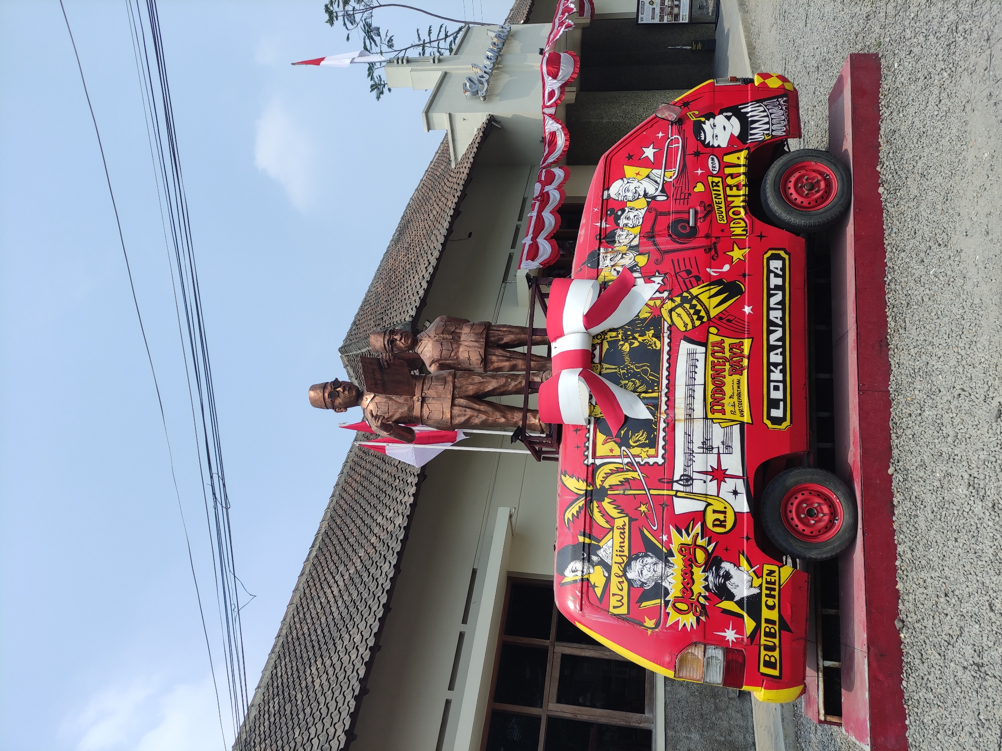 In Surakarta, Indonesia, an outdoor scene features a brightly painted red van with a yellow stripe along its base. The van is adorned with various drawings and text, including depictions of people and musical notes. A large red and white bow is affixed to the top of the van. Two bronze statues of men, one taller and one shorter, stand atop the van. The taller statue holds a book, and both appear to be dressed in formal attire. Two Indonesian flags, one red and white and another red and white with a white motif, are positioned behind the statues. The van is displayed on a red platform, which is surrounded by a gravel-covered ground. In the background, a building with a tiled roof and white walls is visible. Decorative red and white bunting hangs from the building's awning. The sky above is clear and blue with scattered clouds. The overall impression is a festive display, likely related to a cultural or historical event.