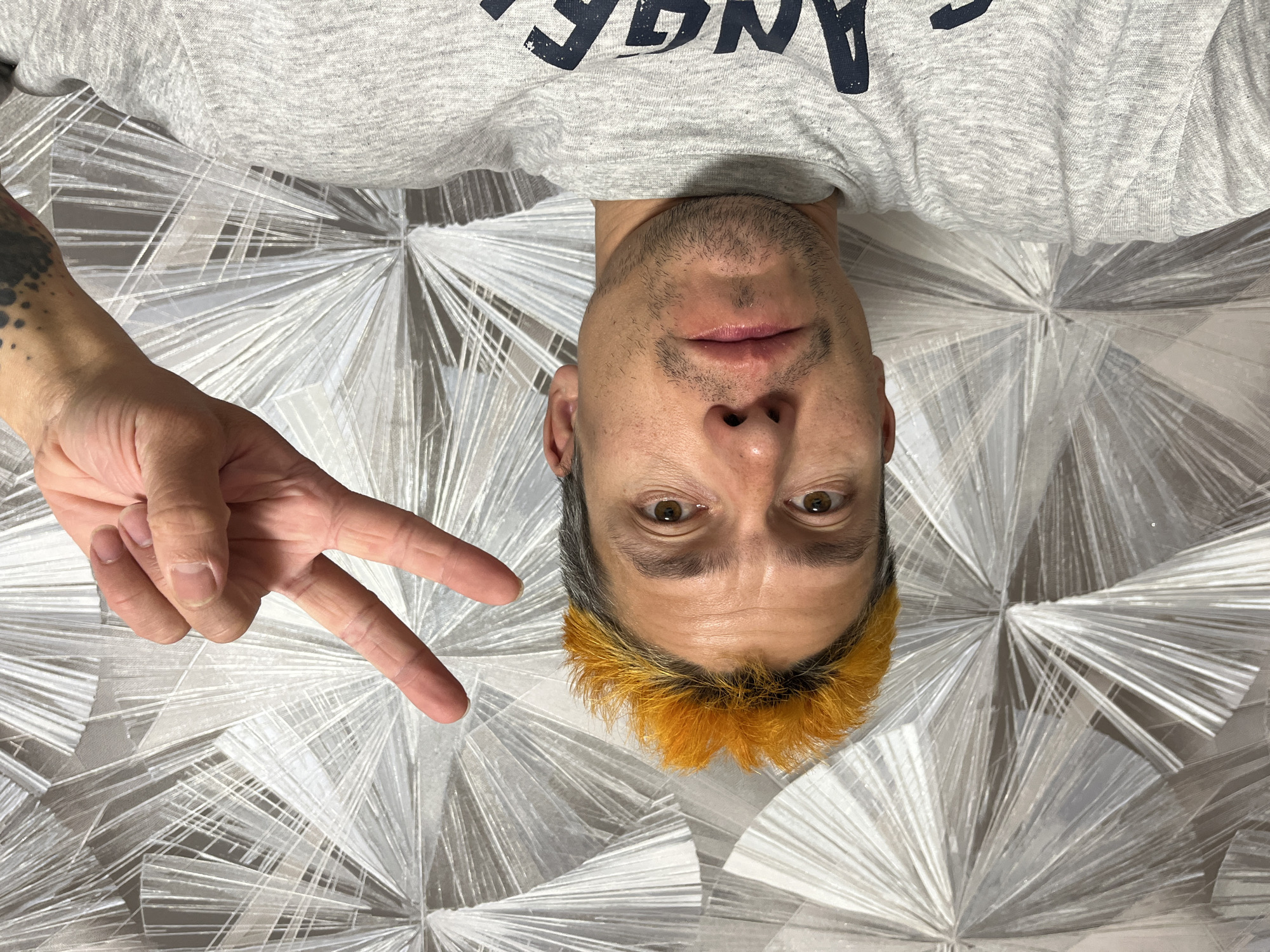 A man lies upside down on a bed with a white and grey patterned comforter. He is wearing a grey shirt with the word "ANGE" partially visible at the top. His hair is grey with orange tips. He is making a peace sign with his left hand. The location cannot be determined from the image, and therefore, it is not possible to confirm the location as Chaikovsky, Russia.