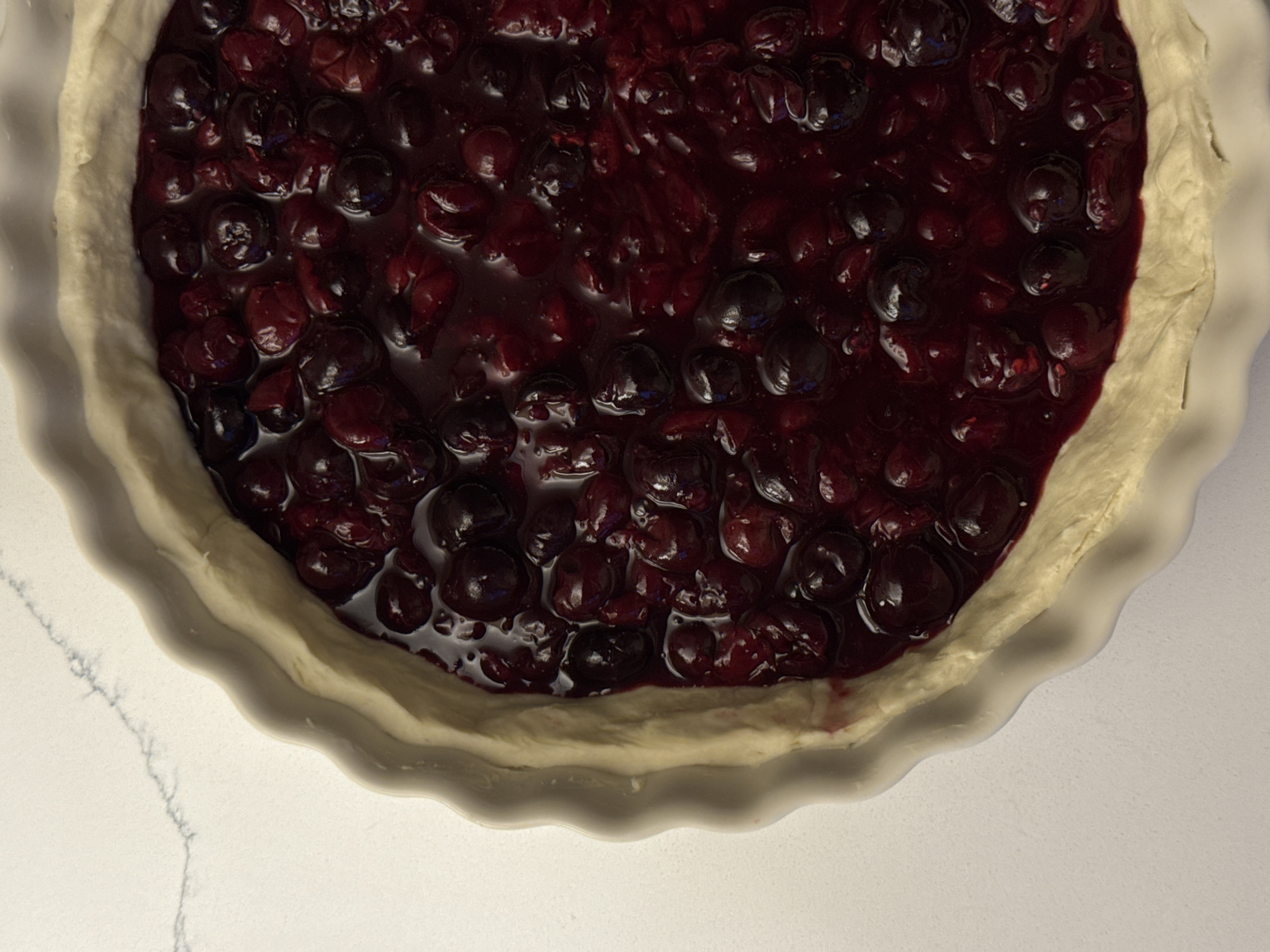 The image shows a cherry pie in a white, fluted pie dish. The filling is a dark red, glossy mixture of cherries. The crust is light in color and appears uncooked. The pie dish sits on a white surface with subtle veining. There is no evidence to suggest a location in the city of Moscow, Russia, or any other location. There are no people, actions, or interactions visible in the image.