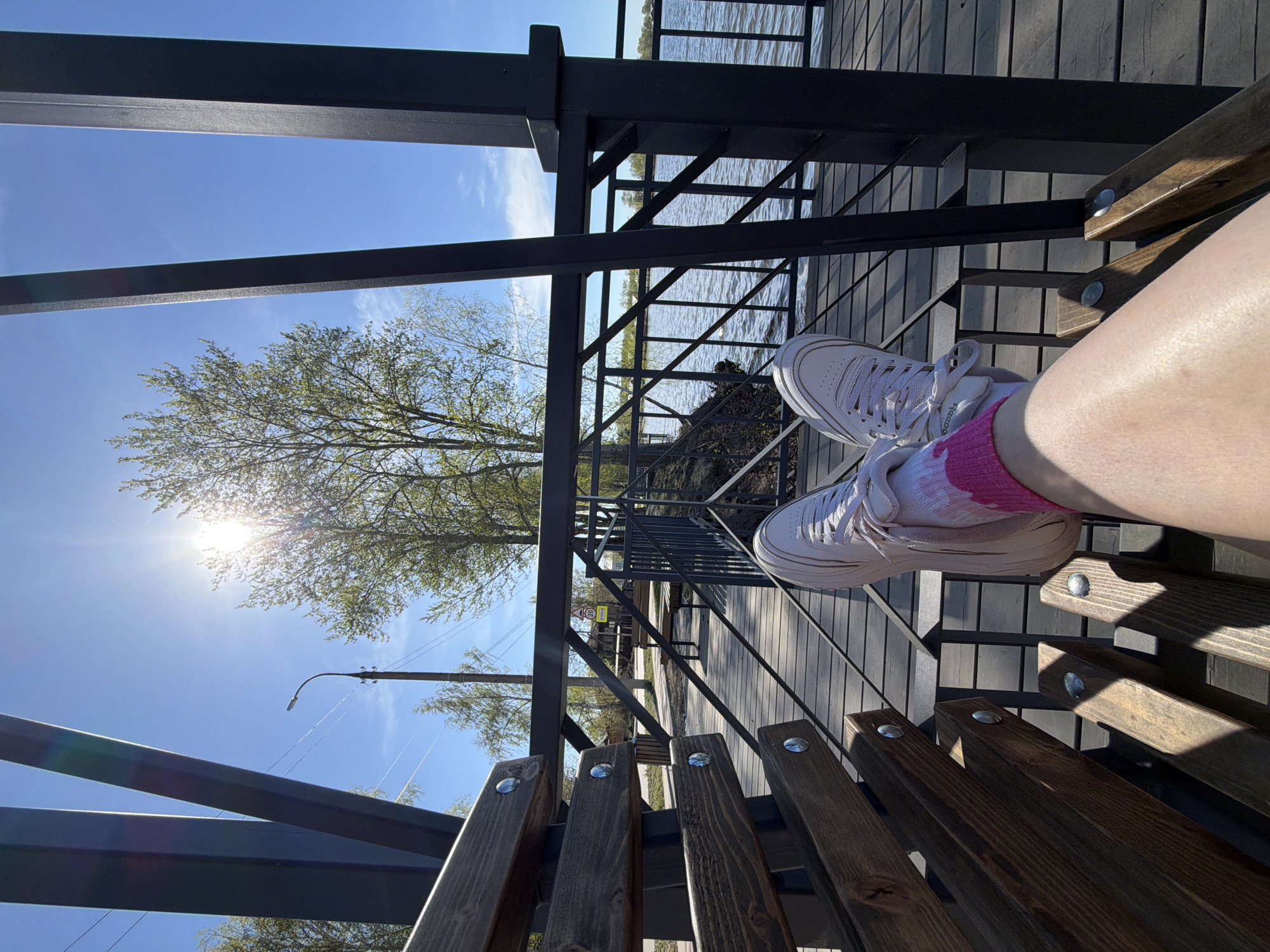 The image depicts a person sitting on a wooden bench with a view of the Votkinsk reservoir in Votkinsk, Russia. The person is wearing white sneakers and pink socks. A metal railing is visible, as well as a tree with bright sunlight shining through its branches. In the background, water can be seen, and the sky is blue. The scene appears to be outdoors on a sunny day.