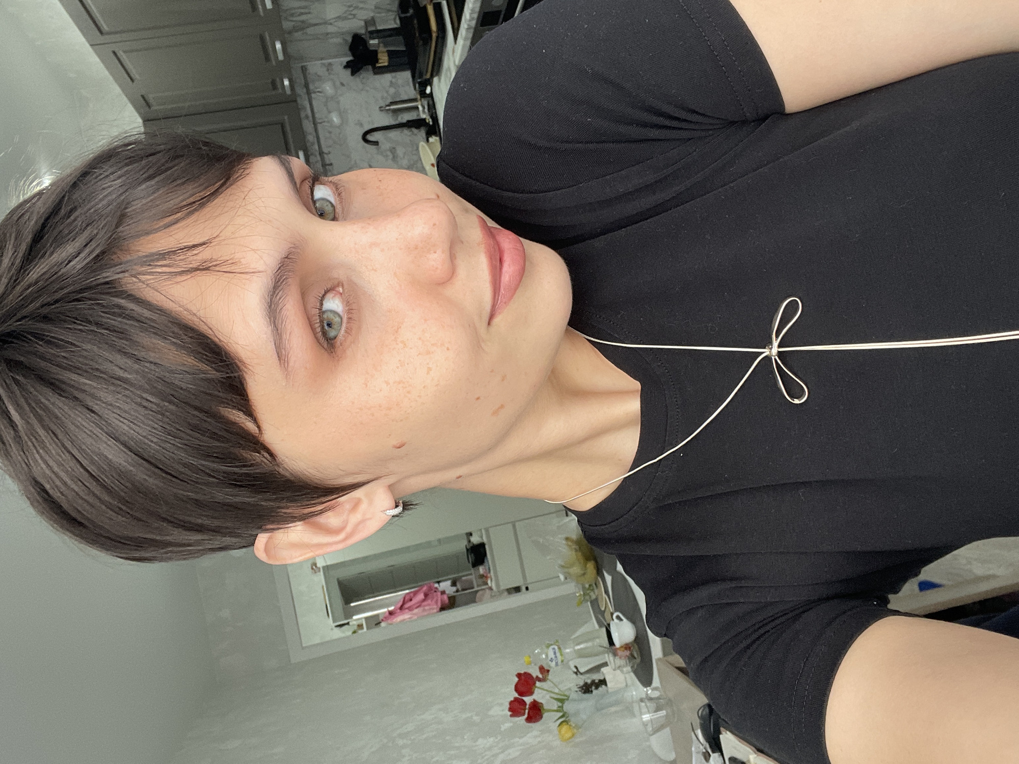 A selfie of a fair-skinned person with short, dark brown hair and light green eyes. Freckles are visible on the face. The person is wearing a black t-shirt with a thin white cord looped around the front. An earring is visible in the left ear. The background appears to be an interior space, possibly a kitchen or dining area, with white or light-colored walls and cabinets. A glimpse of a reflection in a mirror shows additional items, including flowers in a vase on a table. There is no evidence to indicate this photo was taken in Izhevsk, Russia.