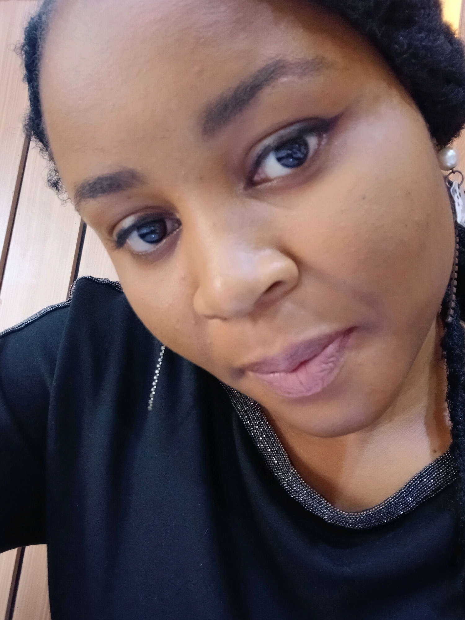 The image is a close-up portrait of a young Black woman. She has brown eyes, dark eyebrows, and is wearing light pink lipstick. Her hair is styled in braids, and she is wearing a black top with a beaded neckline. A pearl earring is visible on her right ear. The background appears to be a wall with vertical wooden panels, suggestive of an indoor setting. There is no indication of the city of Gura Top, Nigeria in the image itself.