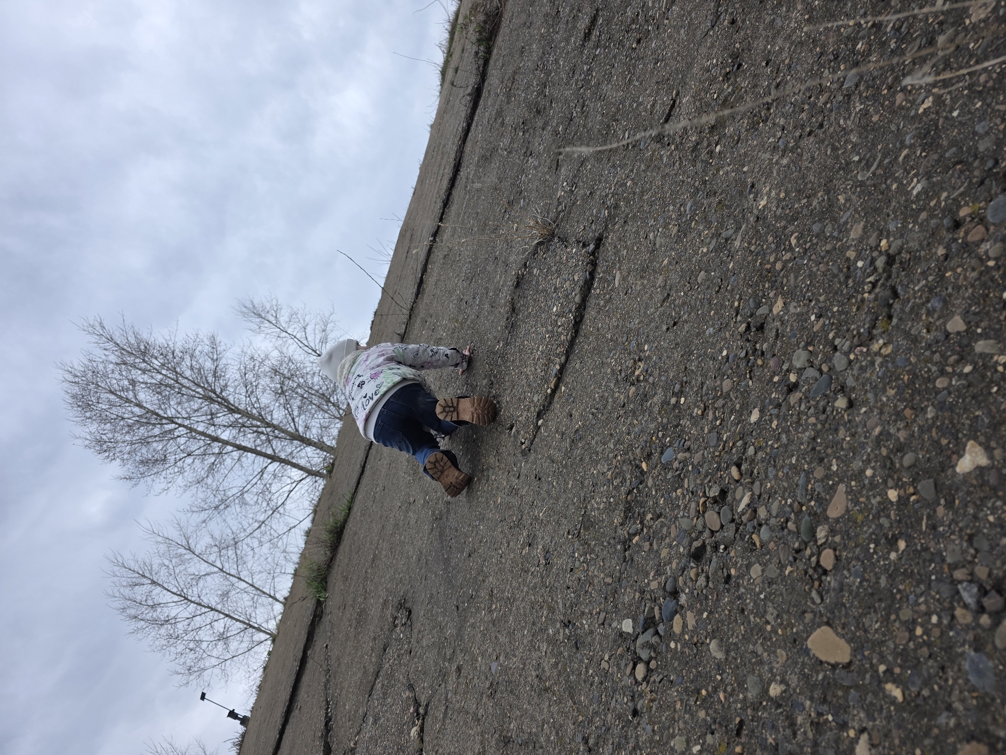 A child, dressed in a white patterned hoodie, dark jeans, and brown boots, is crawling up a weathered, gray concrete slope. The concrete surface is cracked and embedded with small rocks. Sparse, dry grass and a few bare trees are visible near the top edge of the slope against a cloudy sky. The location is Chaikovsky, Russia.
