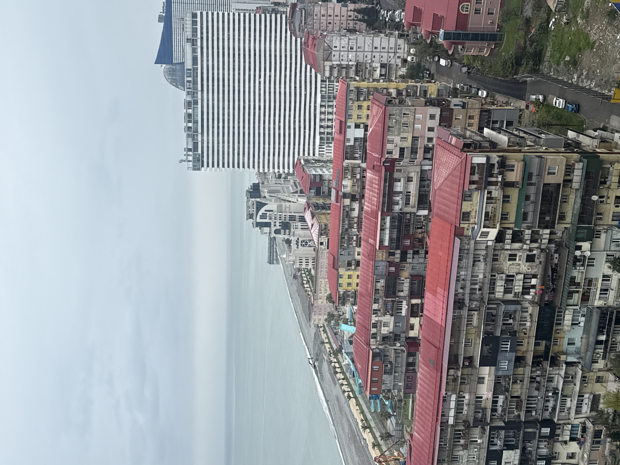 The image captures a view of Batumi, Georgia, from an elevated perspective. The scene is dominated by the Black Sea on the left and a dense urban landscape on the right. A beach is visible along the coast, with a promenade running adjacent. The built environment comprises high-rise buildings, including a modern skyscraper with black and white stripes. Older, Soviet-era apartment blocks with red roofs form the bulk of the housing. A road with a few cars is visible further inland, adjacent to a green area with trees. The weather appears overcast, casting a dull light over the scene.
