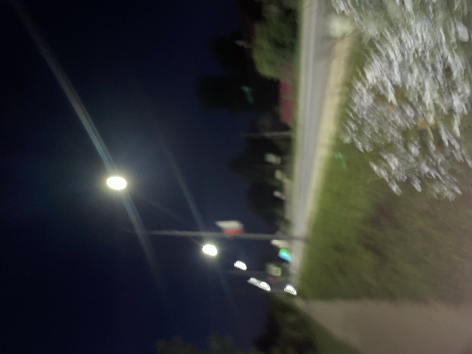 The image captures a blurred nighttime scene along a roadside in the settlement of Krasnye Baki, Russia. A street light illuminates the right side of the frame, casting a bright, diffused glow. To the left of the street light, a flag with red and white sections is visible, possibly indicating a national or local symbol. A sidewalk or path runs along the left edge of the image, bordered by grass and vegetation. Beyond the sidewalk, a road stretches into the distance, with faint lights and shapes suggesting buildings and trees on the other side. The overall impression is one of a quiet, potentially suburban or rural setting at night, with artificial lighting providing the primary source of illumination. The blurriness of the image suggests it may have been taken with a moving camera or in low-light conditions.