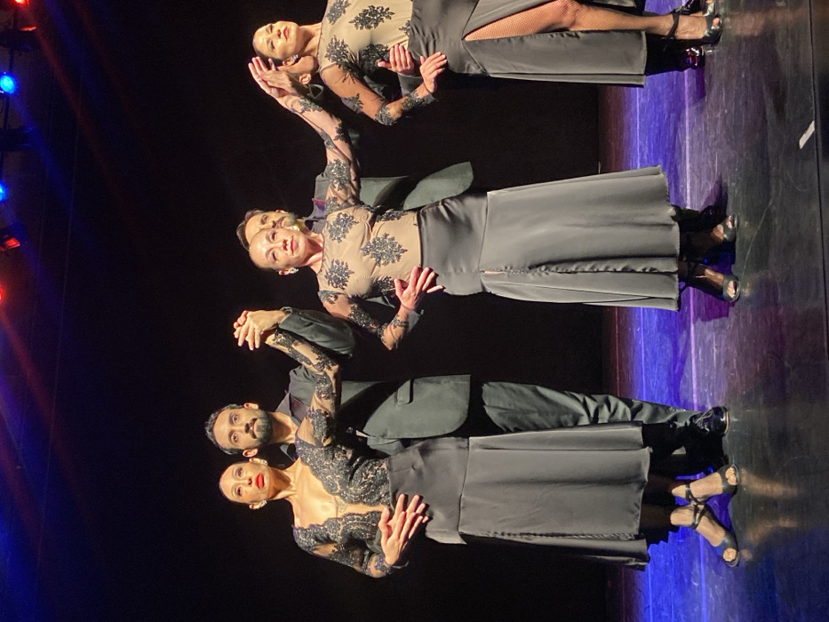 The image displays five individuals, comprising three women and two men, performing on a stage within a performance venue in Buenos Aires, Argentina. They are arranged in a horizontal line across the foreground, against a dark background illuminated by overhead stage lights that cast blue and red hues onto the reflective dark stage floor.
From left to right: A woman, with dark hair pulled back, wears a nude-toned, long-sleeved top adorned with black lace patterning, and a dark grey full-length skirt. Her right hand is clasped with the left hand of the man positioned directly behind her. This man, bearded and dressed in a dark suit with a dark shirt, has his right arm around the woman's waist. Both look forward.
In the center, a second woman, dressed identically in a nude-toned, black-laced top and dark grey skirt with a thigh-high right-side slit, poses with her left hand on her hip and her right arm extended horizontally to her right, palm open. Behind her, a second man in a dark suit is partially visible.
To the right, a third woman wears the same nude-toned, black-laced top and a dark grey skirt with a prominent thigh-high right-side slit, revealing a leg covered in fishnet stockings. Her right hand rests on her hip, and her left arm is extended horizontally to her left, palm open, appearing to connect with the central woman's hand. She looks toward the right.
All visible women wear black open-toed dance shoes with ankle straps and a low heel. All individuals maintain focused, neutral expressions, indicative of a synchronized dance performance.