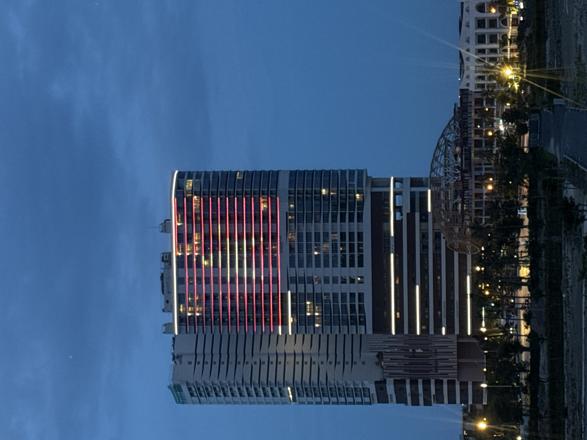 This is a nighttime, vertical, medium-shot photograph of a brightly lit, modern skyscraper in the city of Nha Trang, Vietnam. The skyscraper is the primary subject and is illuminated with red and white lights on its upper floors, forming horizontal bands. The building has a sleek, contemporary design with visible windows and balconies. To the left of the main skyscraper is a slightly shorter, also modern building with a more angular facade. In the foreground, there is a dark, somewhat undeveloped area with scattered trees and streetlights. Further in the background, to the right of the skyscraper, are other buildings with illuminated windows and a large, dome-like structure that appears to be under construction or is a unique architectural feature. The sky is a deep twilight blue, with hints of clouds and lens flare from the lights. The overall impression is one of a vibrant, modern urban landscape at dusk or night.