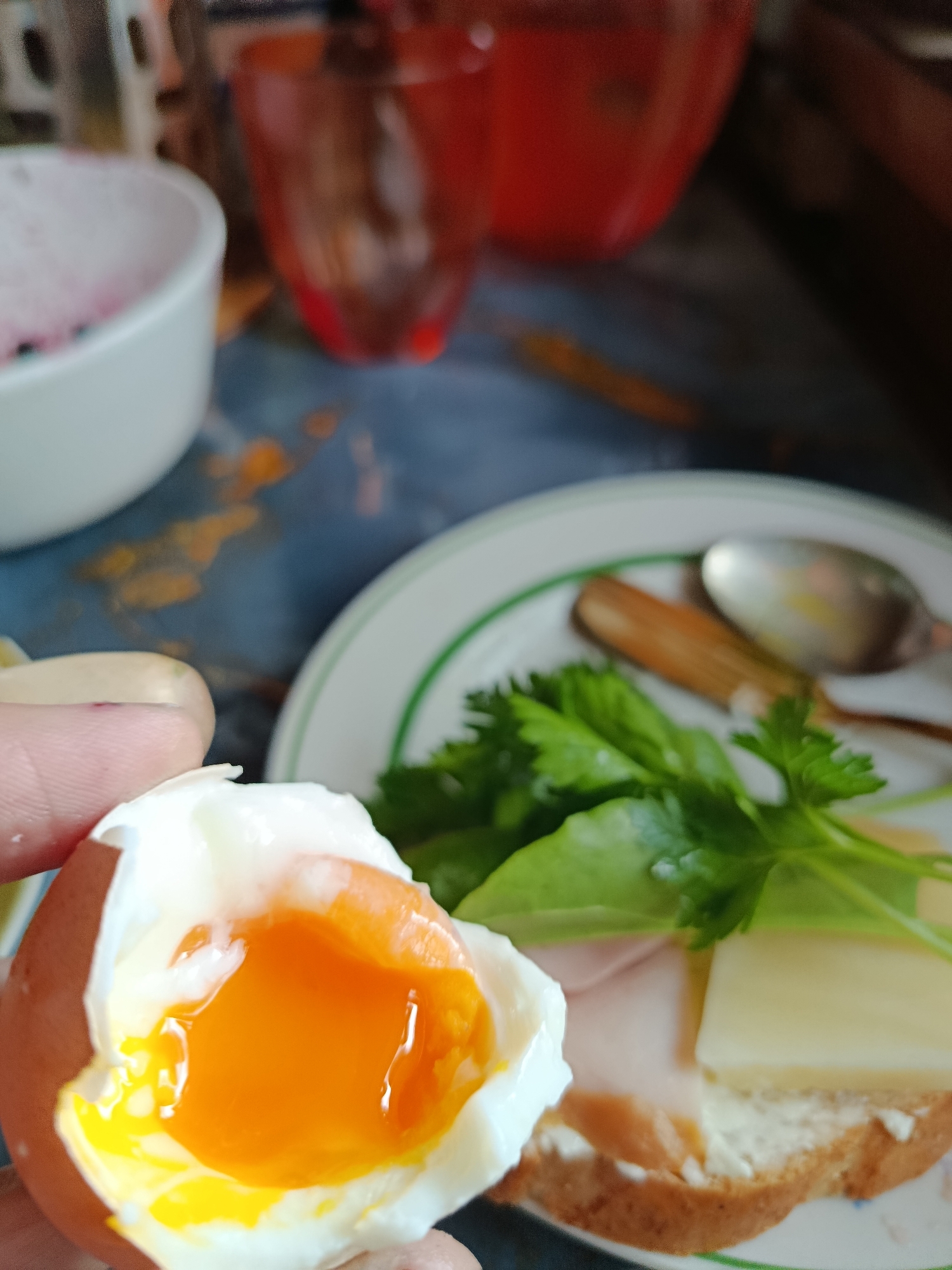 A close-up shot captures a hand holding a cracked soft-boiled egg with a vibrant orange yolk. In the background, a plate contains a sandwich with ham, cheese, and greens. To the left, a white bowl and a blurry red glass are visible. The scene is set against a dark, textured surface, possibly a table. The lighting is soft, highlighting the textures of the food. The image lacks any identifiable landmarks or contextual cues to determine a specific location within a city.