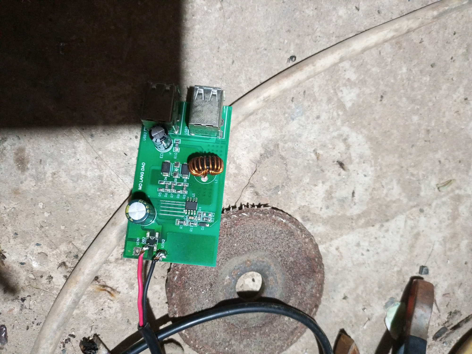 The image shows a green circuit board with two USB ports, a copper coil inductor, a capacitor, and various other electronic components. Red and black wires are connected to the circuit board. The circuit board is placed on a rough, concrete-like surface, possibly outdoors. In the background, a cream-colored cylindrical object, possibly a hose or cable, runs diagonally across the frame. To the right of the circuit board, a rusty, circular metal object with a hole in the center is visible, resembling a cutting wheel. The lighting suggests an indoor or shaded outdoor environment with a shadow cast from the top left. The overall scene appears to be a cluttered, functional workspace, possibly associated with electronic repair or DIY projects. There are no people or clear indications of a specific location within the city of Keling Panggau, Indonesia.