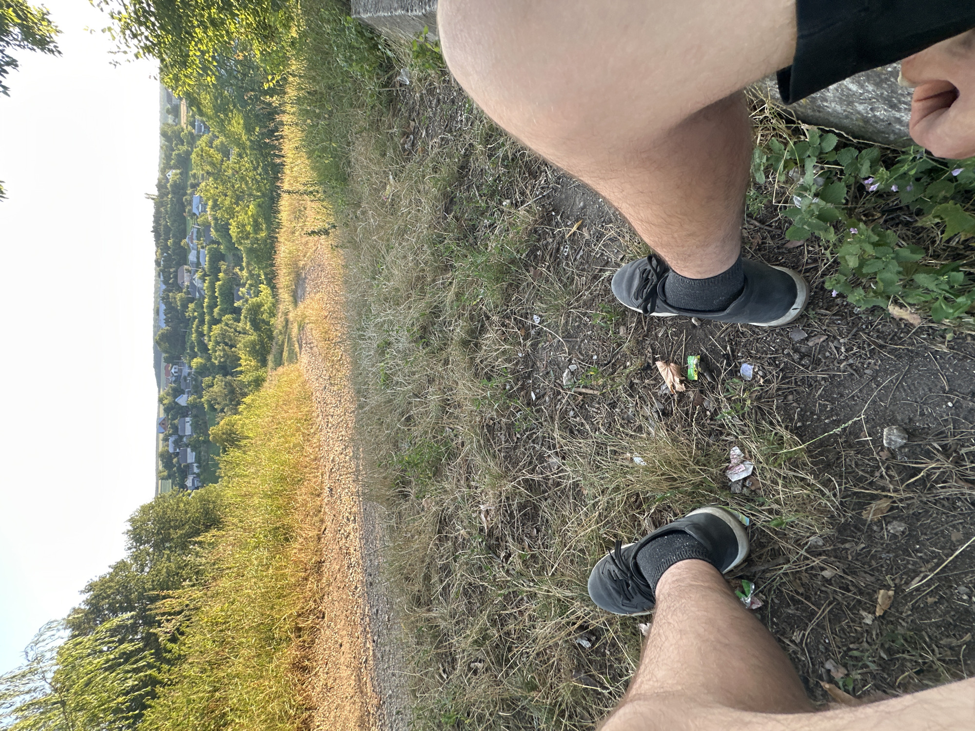 The image shows a first-person perspective of a person sitting outdoors with a view of a small village. The person is wearing dark sneakers and grey socks, and their legs are visible. The ground is covered with dry grass, dirt, and some scattered litter. In the background, a gently sloping hill leads to a village with small houses, surrounded by trees and green vegetation. The sky is clear and bright, suggesting it is daytime, likely late afternoon given the warm light. The location is identified as Makiv, Ukraine.