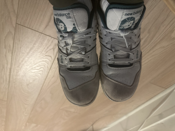 A top-down view showcases a pair of New Balance 550 sneakers. They feature a color scheme of white, gray, and dark green accents. The white leather body displays wear and creasing, contrasted by gray suede toe caps. Laces are neatly tied, and the "550" insignia is visible near the laces. The New Balance logo, incorporating a basketball design, is prominently displayed on the tongue of each shoe. The footwear rests on a light-colored wooden floor.