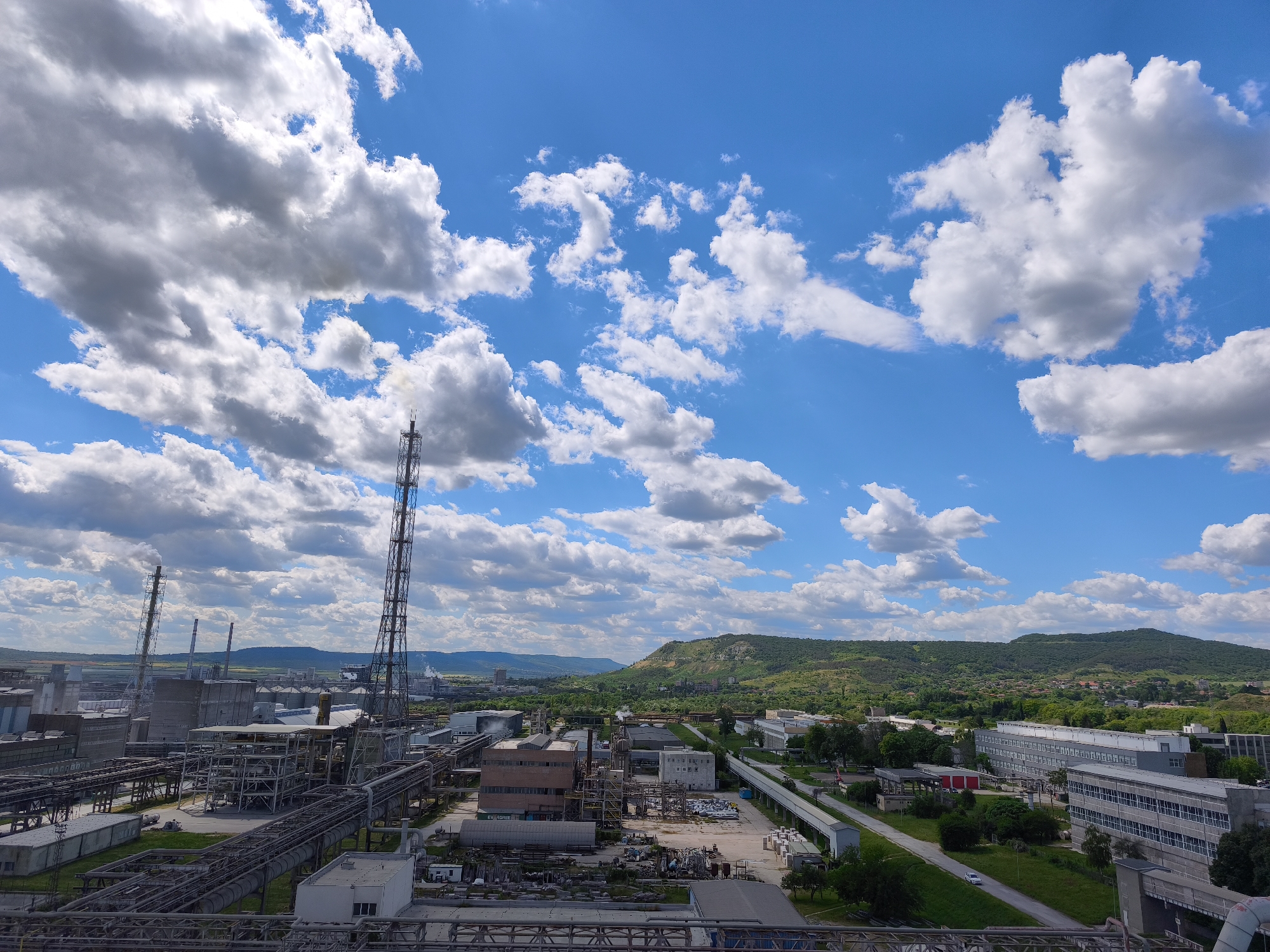 The image shows an industrial area, possibly a chemical plant or factory complex, under a bright blue sky with scattered white clouds. Multiple metal structures, pipes, and buildings are visible, indicative of industrial processing. Tall smokestacks extend upward, one emitting a small plume of smoke. In the background, there are green hills and a distant cityscape. The scene is taken from a high vantage point.